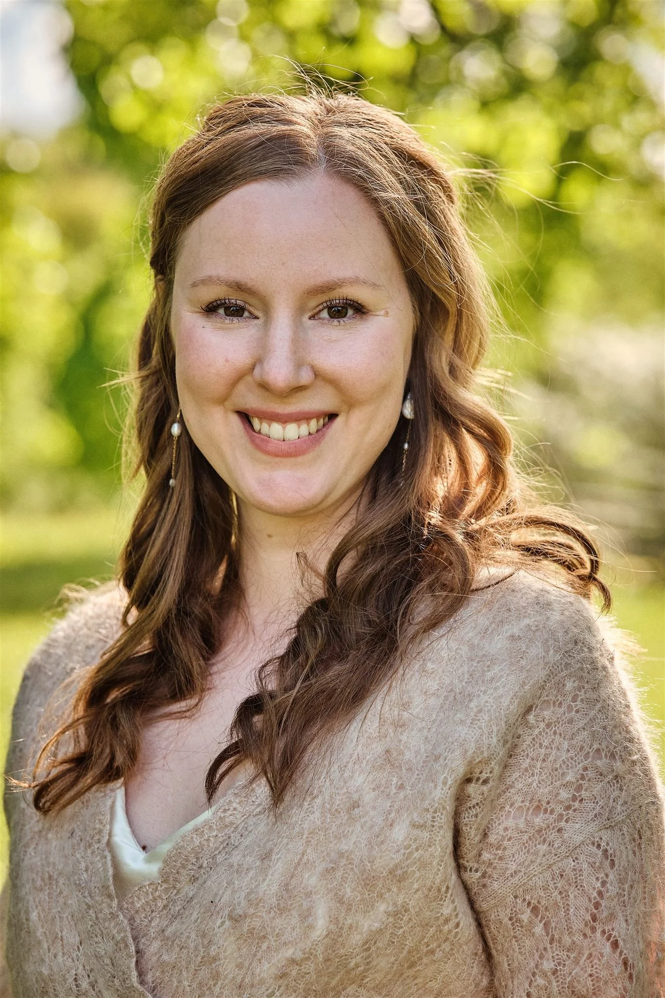 A woman with long, wavy auburn hair, smiling outdoors. She is wearing earrings and a beige lace cardigan, with a white top underneath. The background is green and blurred, indicating a natural environment.