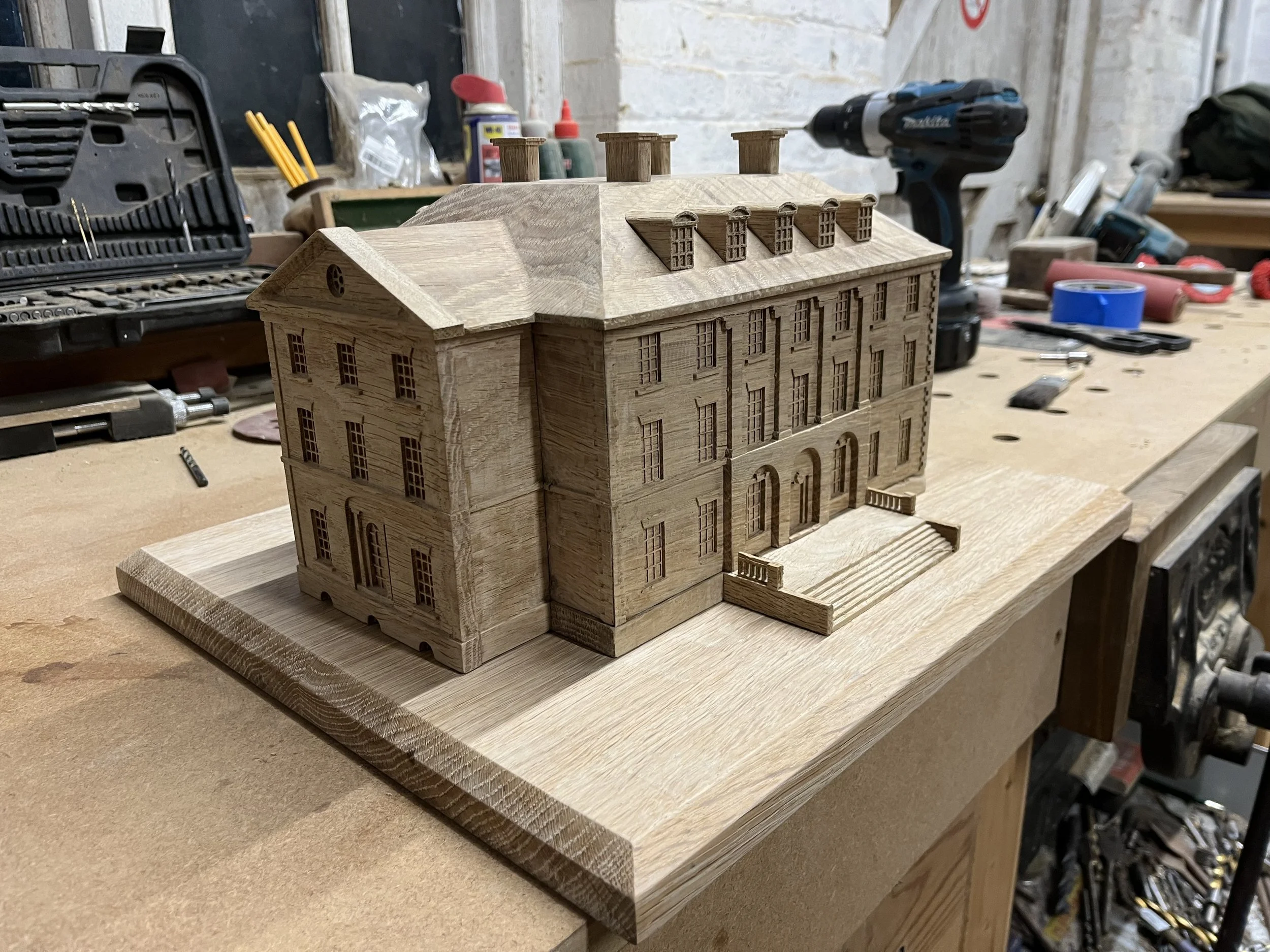 A detailed miniature wooden model of a multi-story building with architectural details, placed on a workbench in a woodworking shop.