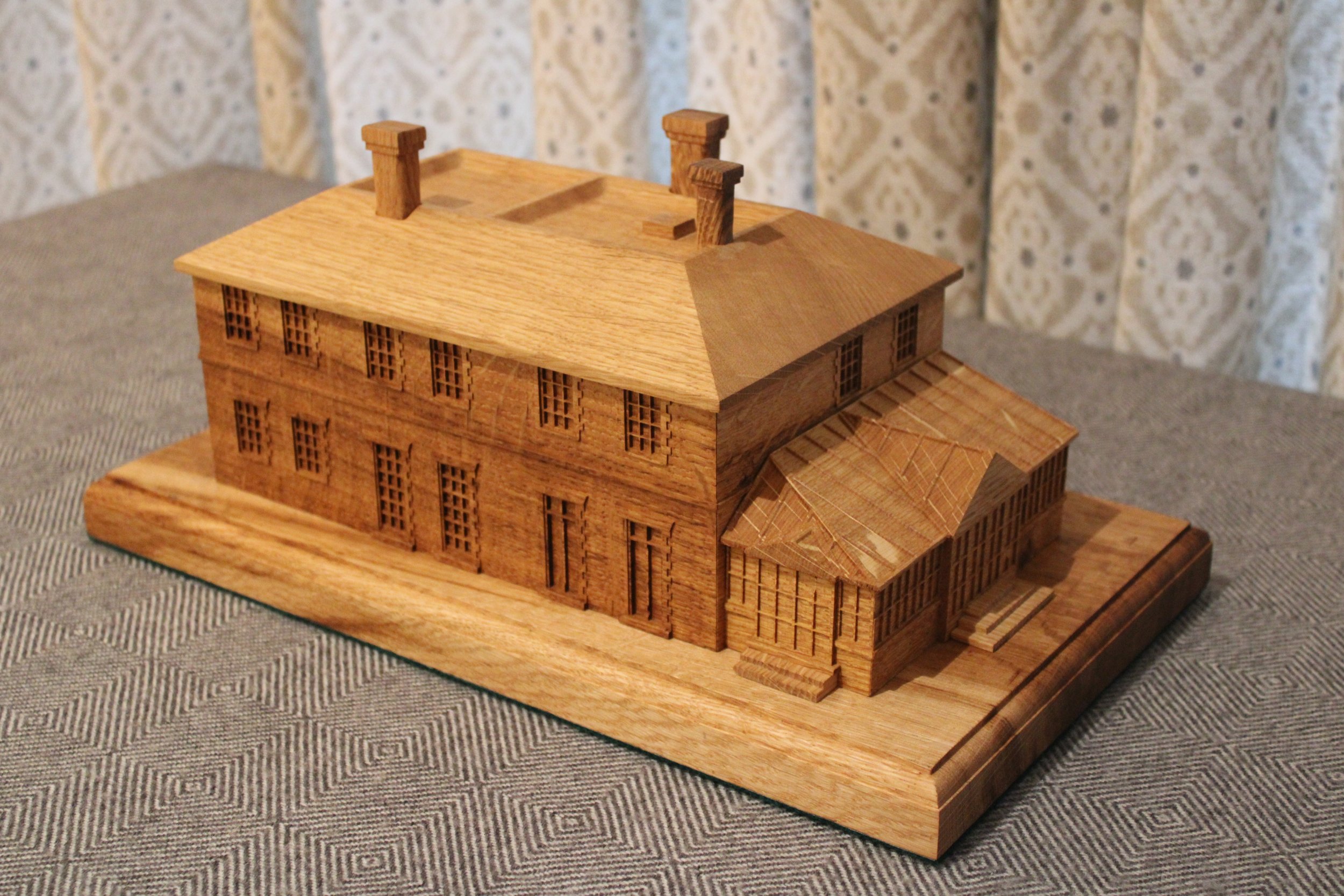 A detailed wooden architectural model of a large building with multiple stories, a sloped roof, and chimneys, placed on a wooden base.