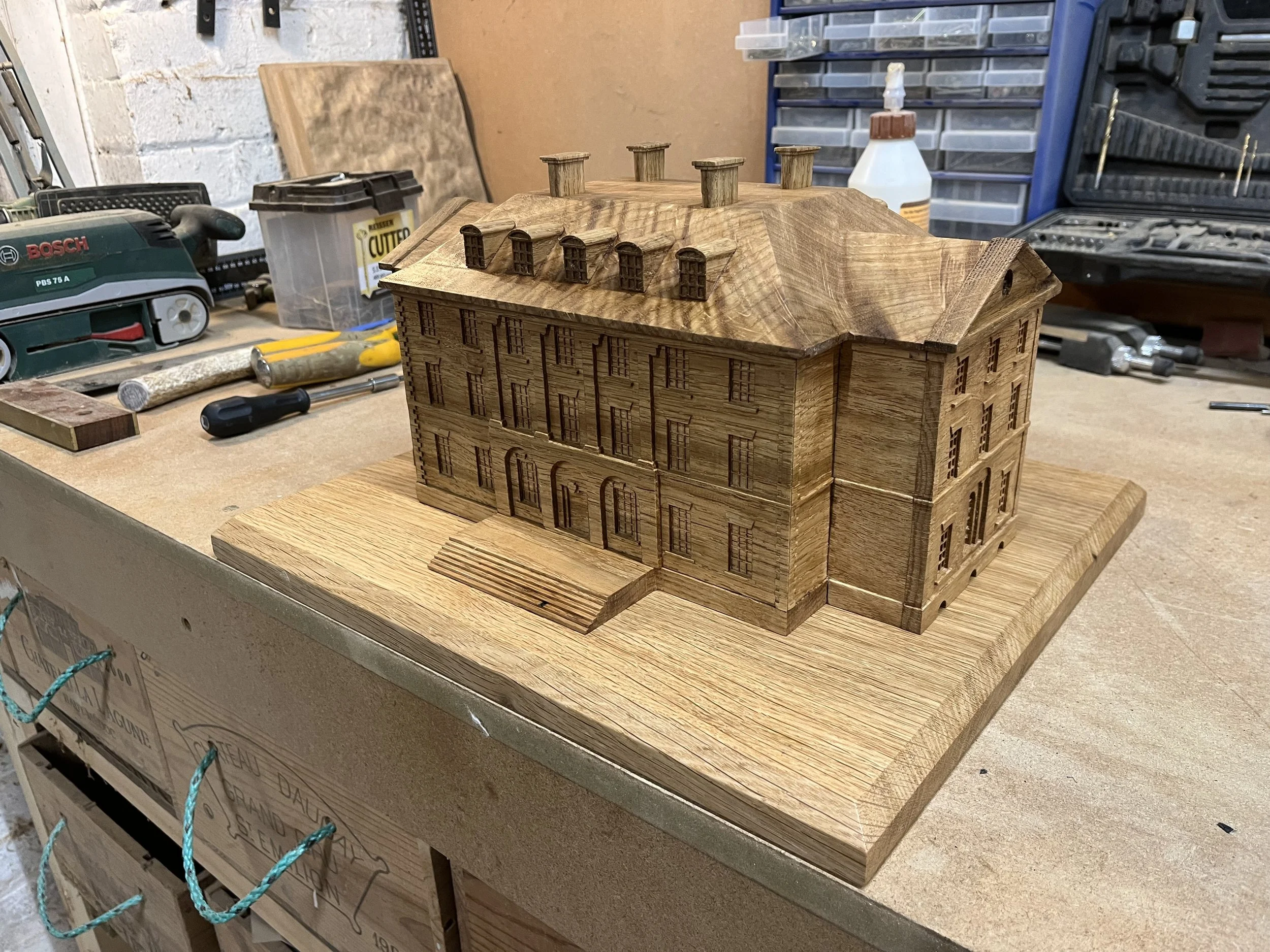 Wooden architectural model of a large three-story building with multiple windows, stairs, and a sloped roof, on a workbench in a woodworking shop.