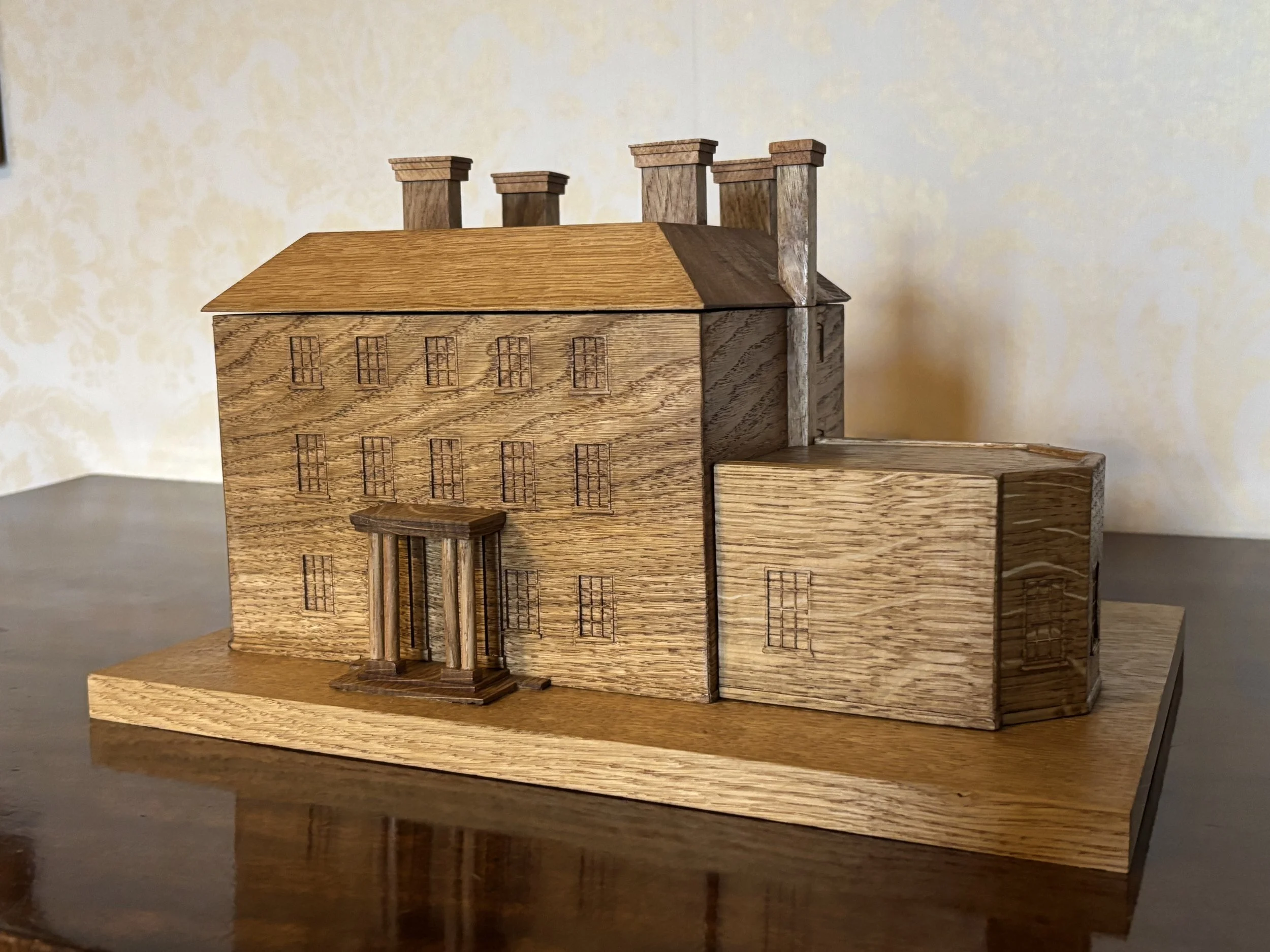 Wooden model of a building with a main structure and an attached extension, featuring multiple chimney stacks on the roof.