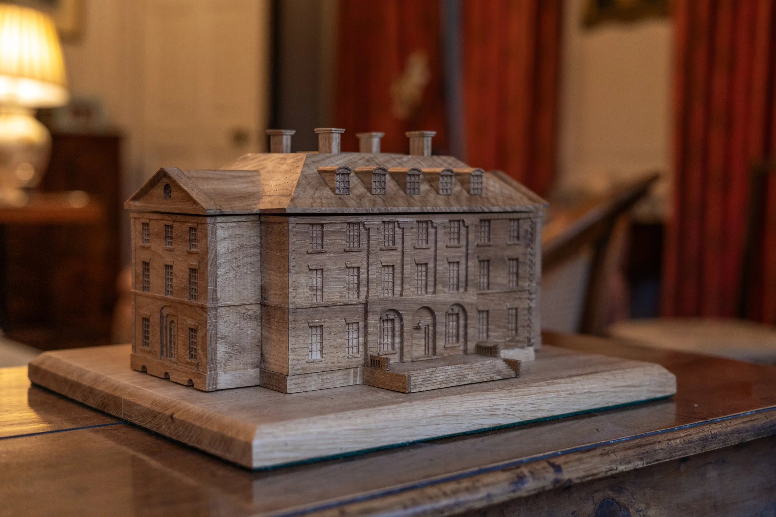 Wooden model of a multi-story building with detailed windows, doors, and roof chimneys, placed on a wooden surface in a warmly lit room.