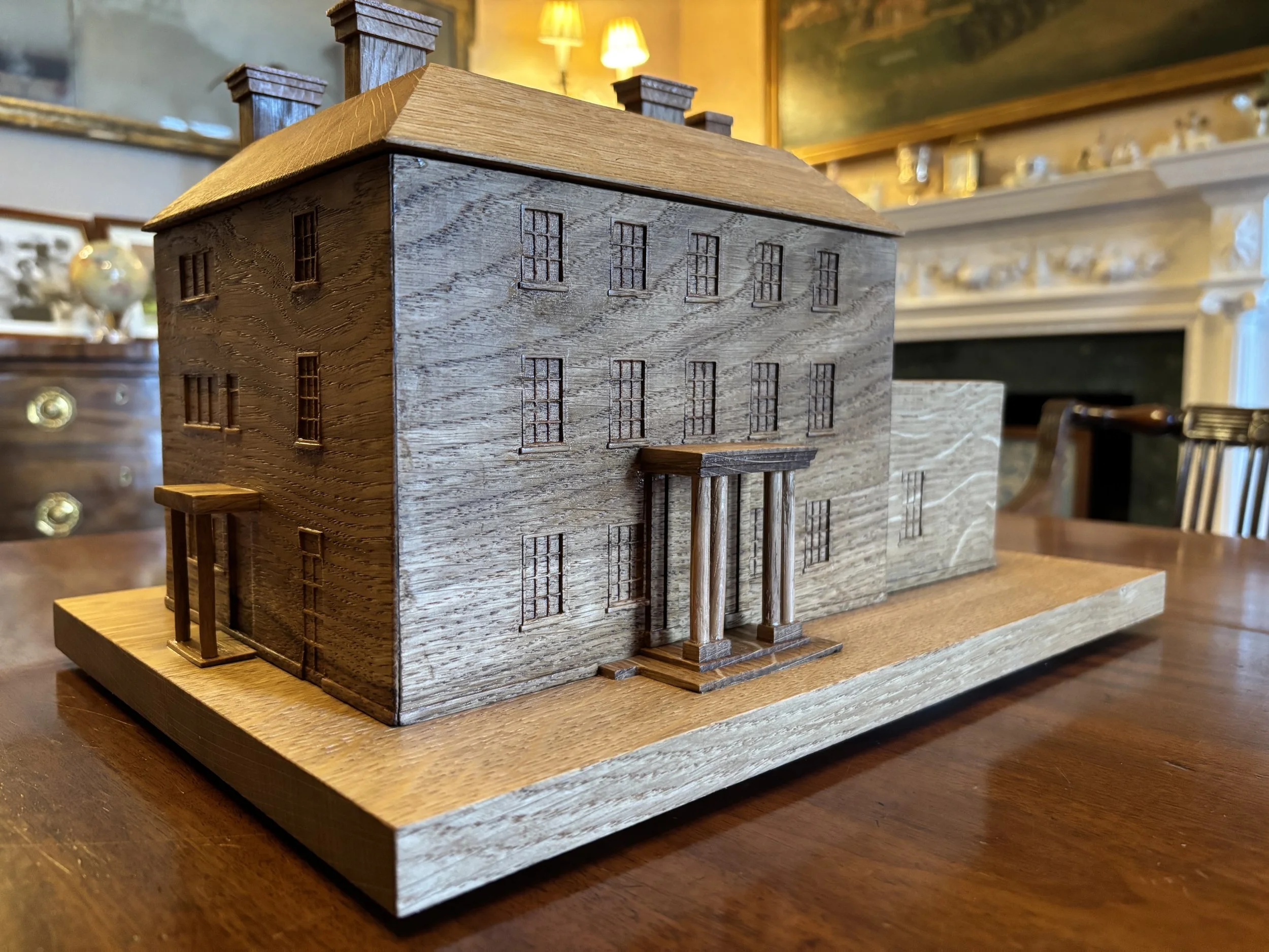 Wooden miniature house model with detailed windows, door, and porch, placed on a wooden table in a room with antique furniture and a fireplace.