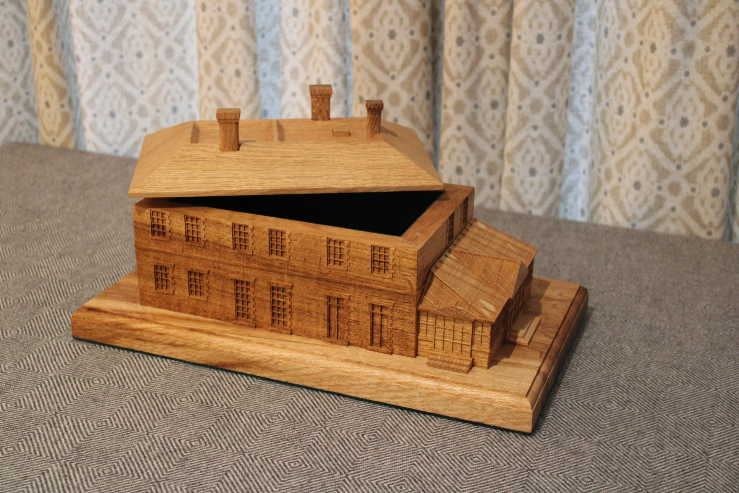 Wooden architectural model of a house with open roof and chimney on a wooden base, placed on a fabric-covered surface.