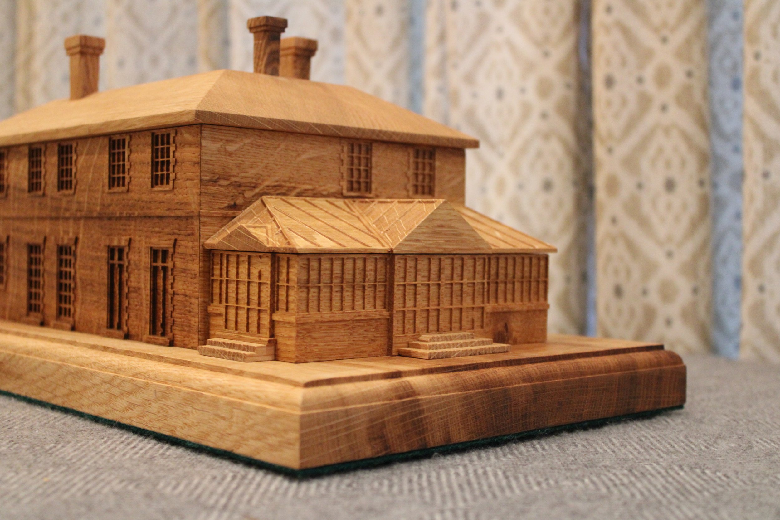 A detailed wooden model of a large, multi-story building with multiple windows and steps leading to the entrance, placed on a gray surface with a patterned curtain in the background.