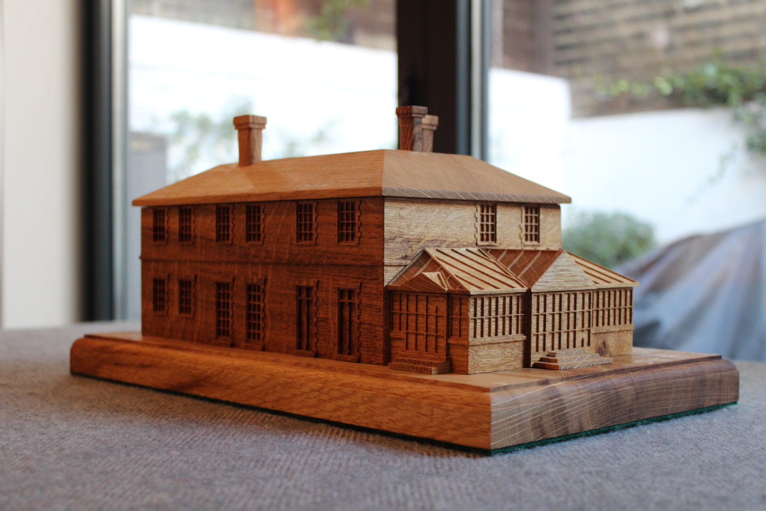 A detailed wooden architectural model of a large, historic-style building with multiple chimneys and intricate window and roof designs, placed on a fabric-covered surface near a large window.