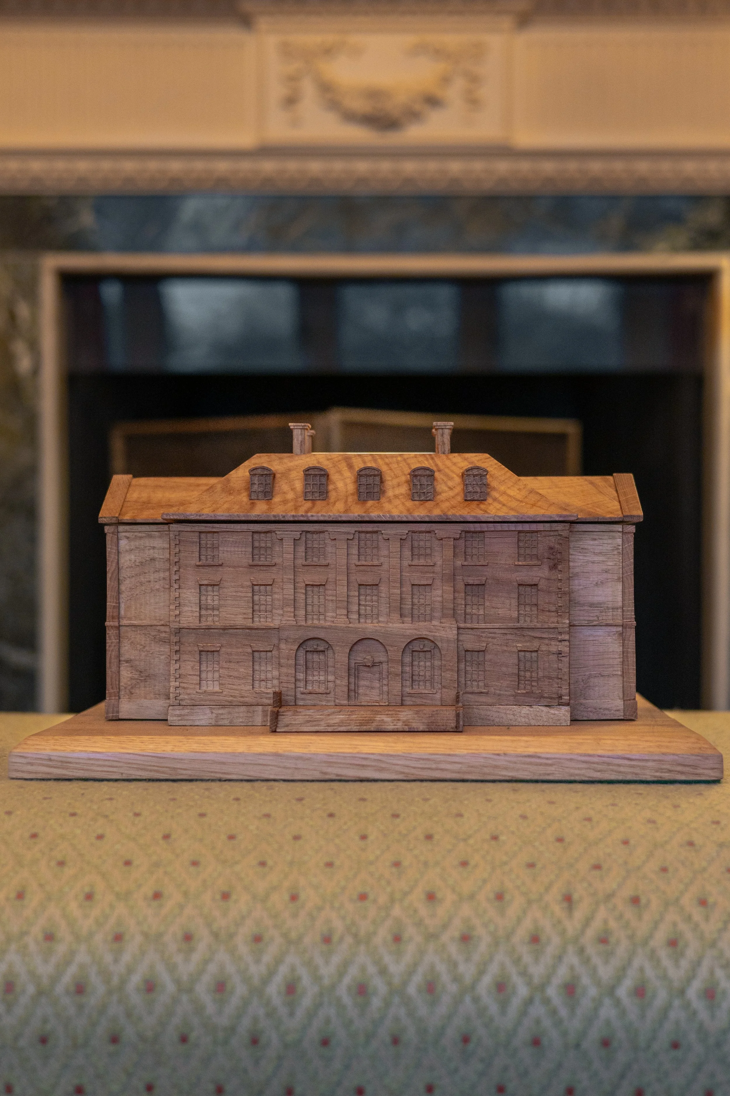 Wooden model of a large classical style building, placed on a table with patterned green fabric, in front of a fireplace.