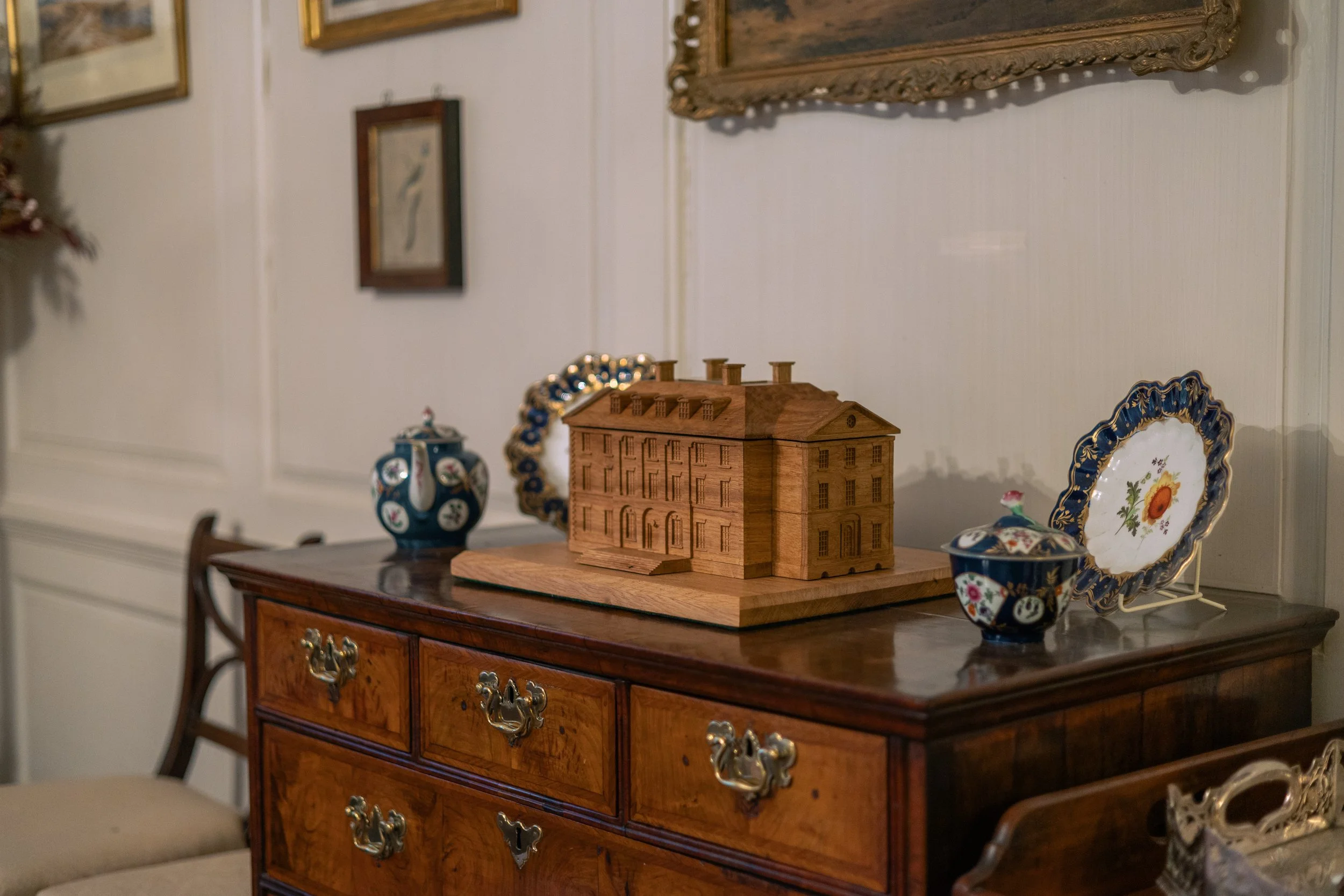 A wooden sideboard with brass handles holds decorative items including a detailed wooden house model, a floral porcelain vase, a ceramic jar with a lid, and a decorative plate on a stand. Behind the sideboard, there is a wall with framed artwork.