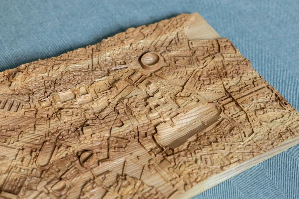Close-up of a wooden carved map or cityscape with detailed buildings and streets, resting on a blue fabric surface.