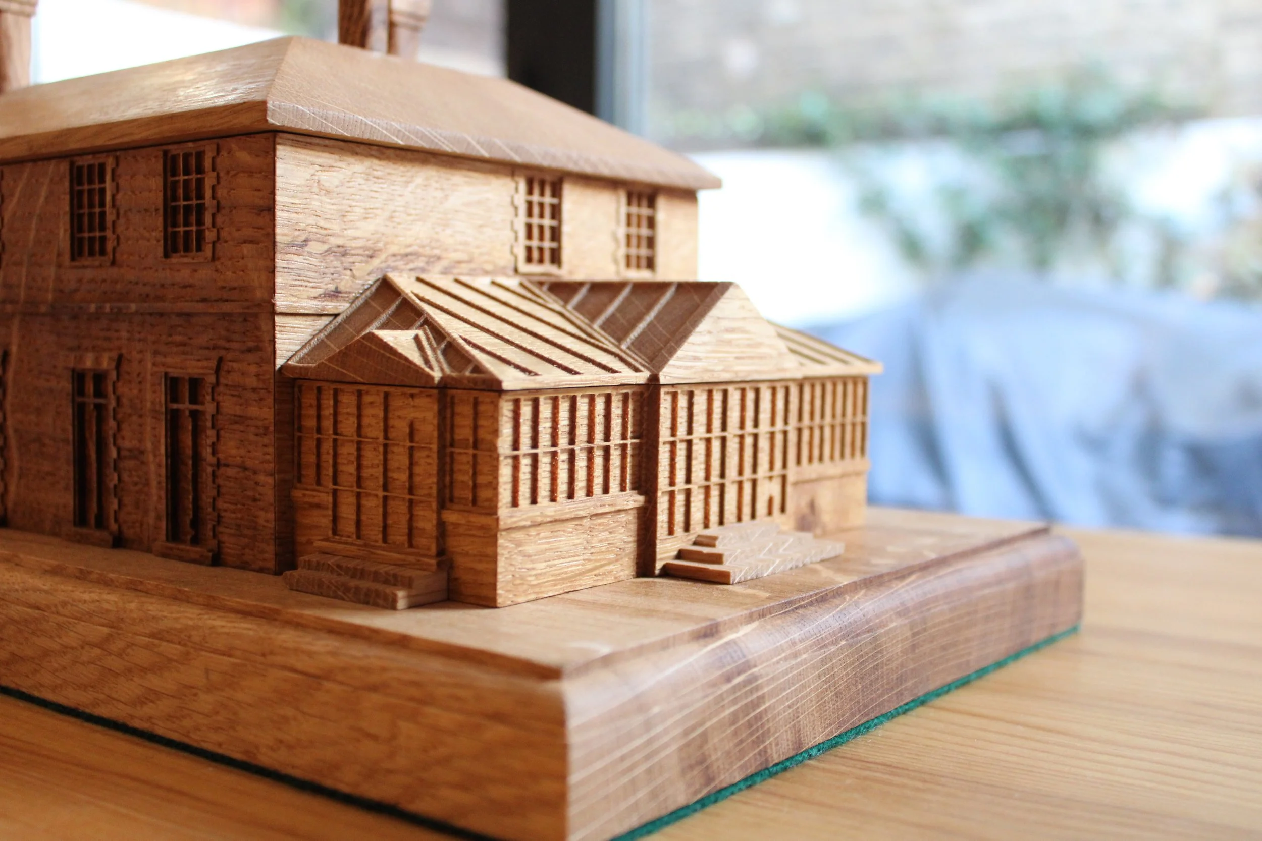 Wooden architectural model of a house with multiple sections and steps, placed on a wooden surface, with a blurred outdoor scene in the background.