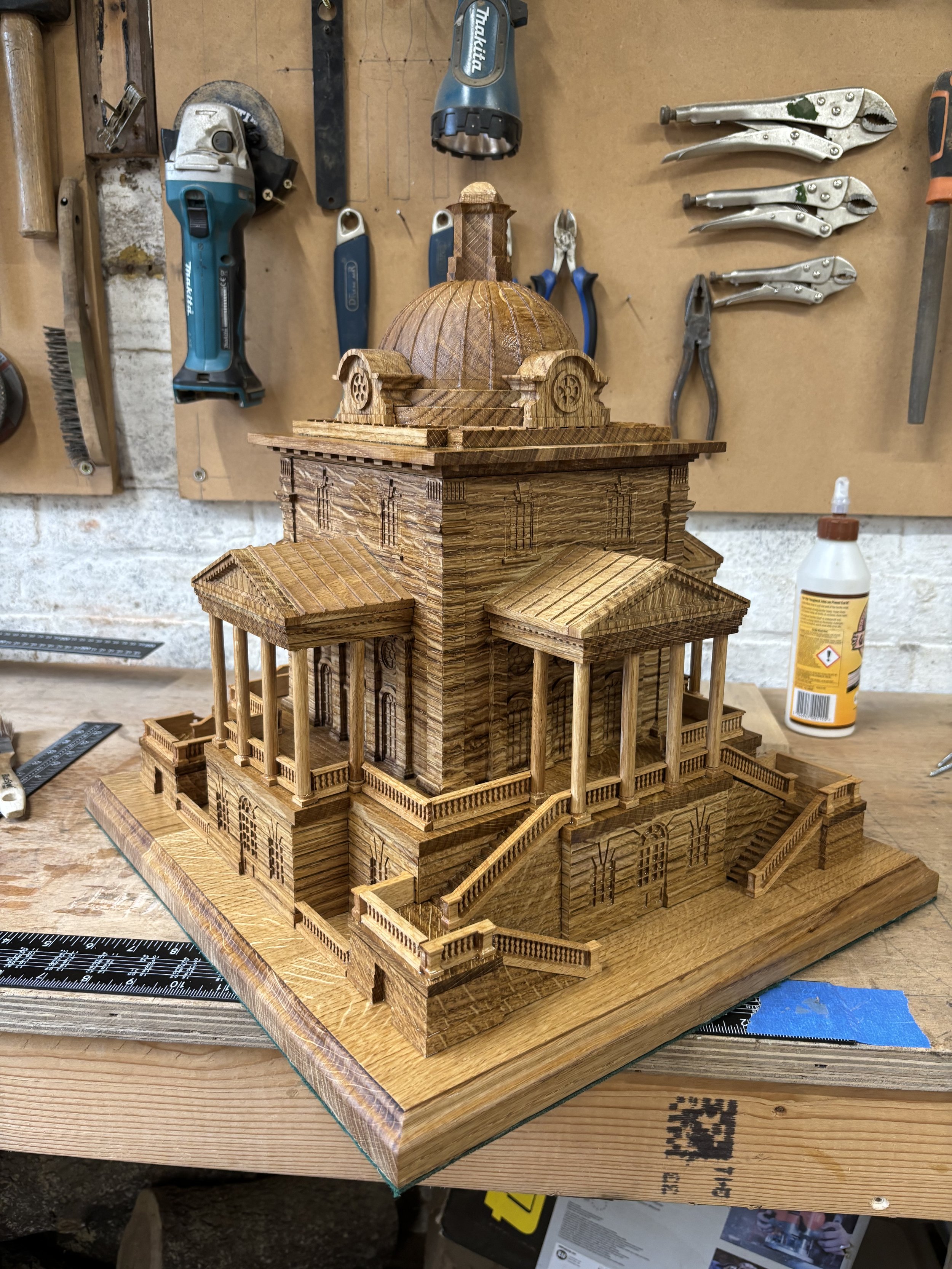 A detailed miniature wooden model of a classical building or temple with columns, stairs, and a dome, placed on a workbench in a workshop with tools hanging on the wall behind.