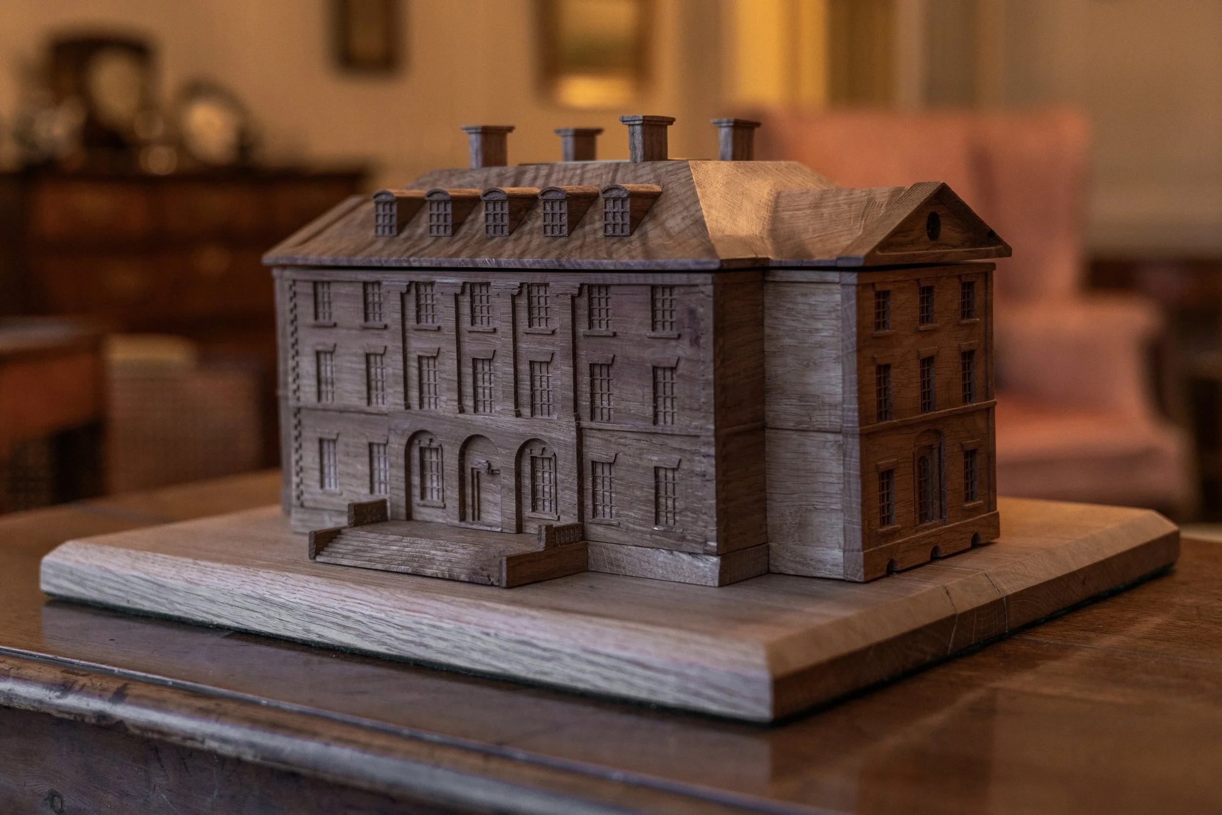 A detailed wooden model of a grand, multi-story building with numerous windows, an arched doorway, and a small staircase, placed on a wooden base indoors.