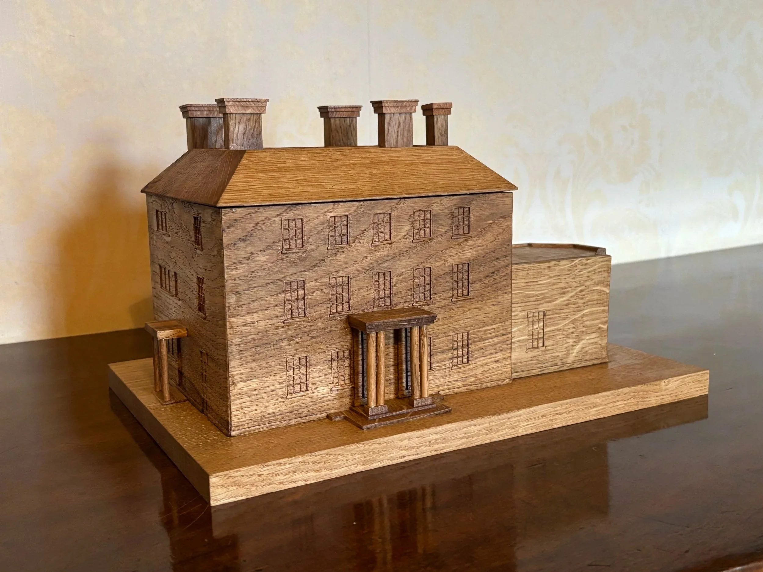 Wooden architectural model of a multi-story building with a rectangular base, multiple windows, a pitched roof, and chimneys, placed on a wooden surface.