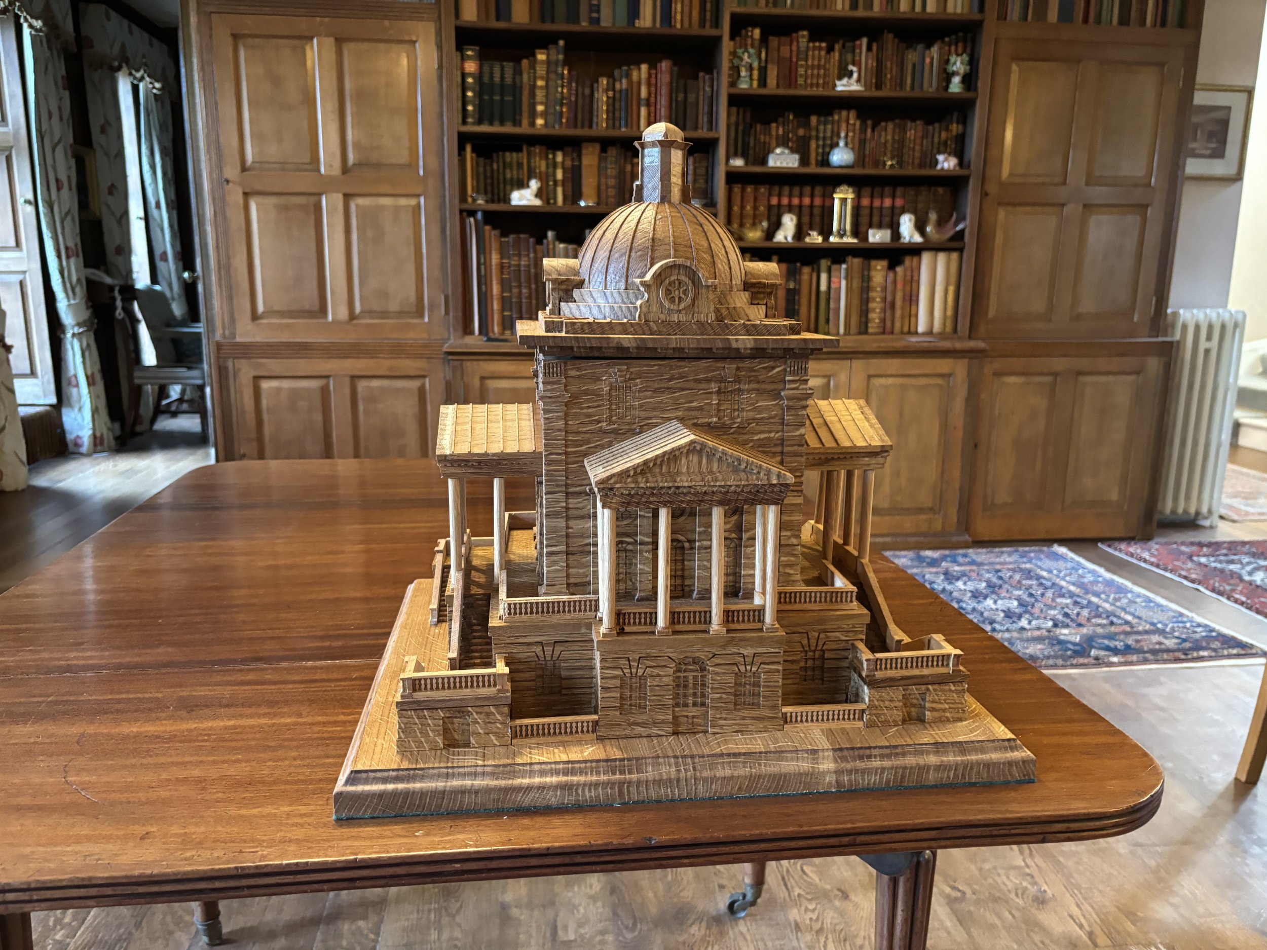 A detailed wooden model of a classical temple with a dome, columns, stairs, and surrounding walls is placed on a wooden table. In the background, there is a large wooden bookcase filled with books and decorative items.