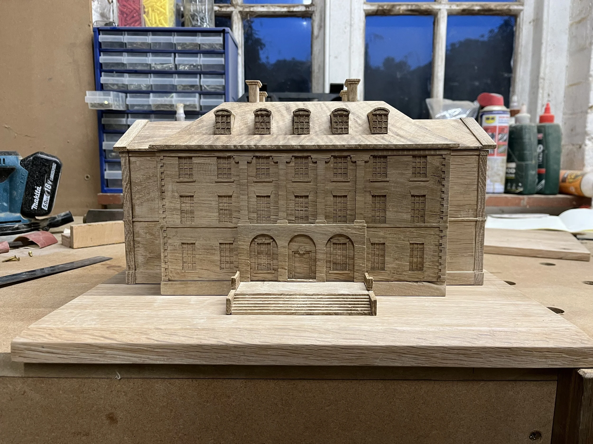 Miniature wooden model of a large, multi-story building with detailed window and door designs, stairs leading to the entrance, and a pitched roof with multiple chimneys, set on a wooden base in a workshop.