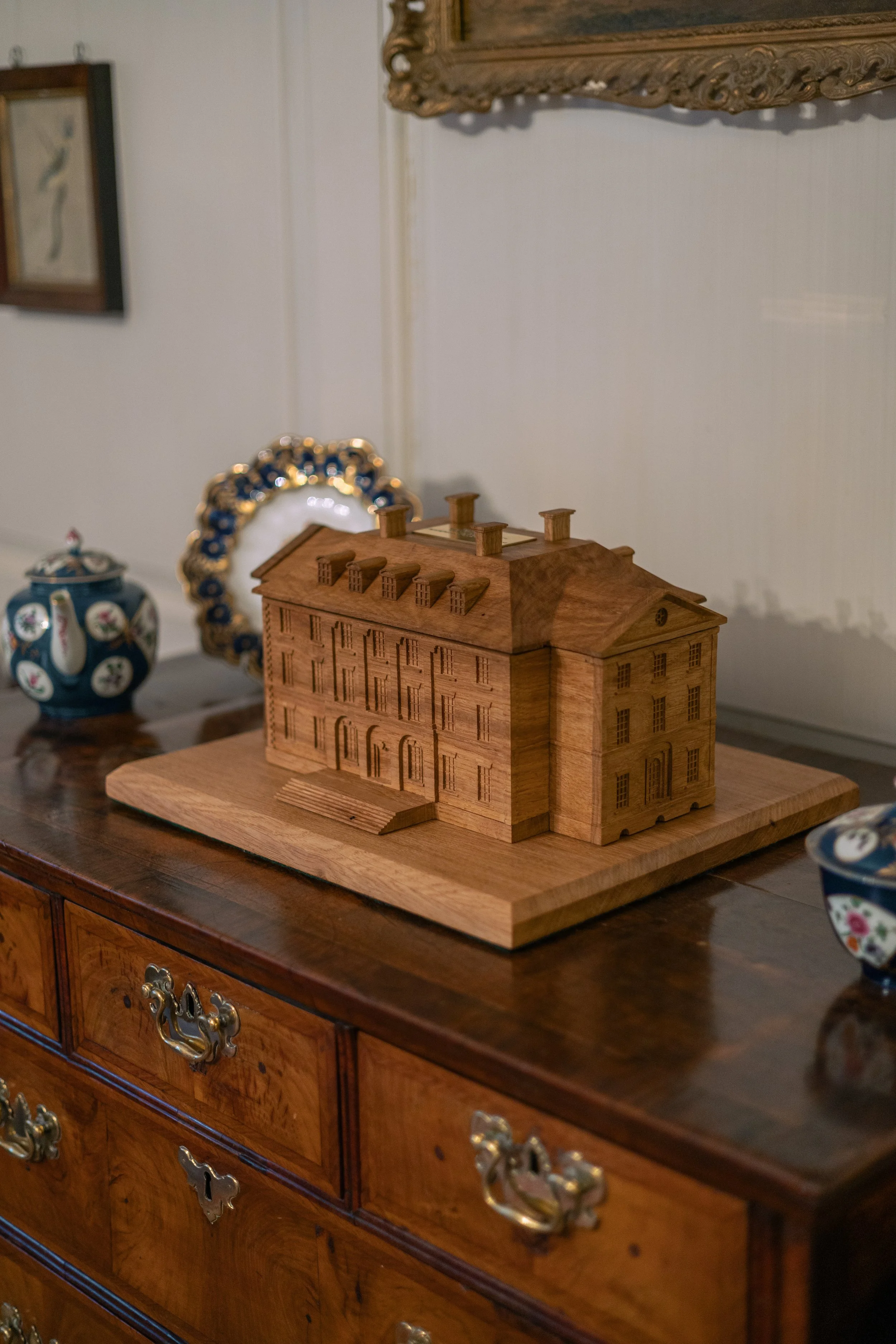 Wooden model of a mansion house on a wooden table, flanked by decorative porcelain teapots, in a room with framed artwork and ornate mirror.