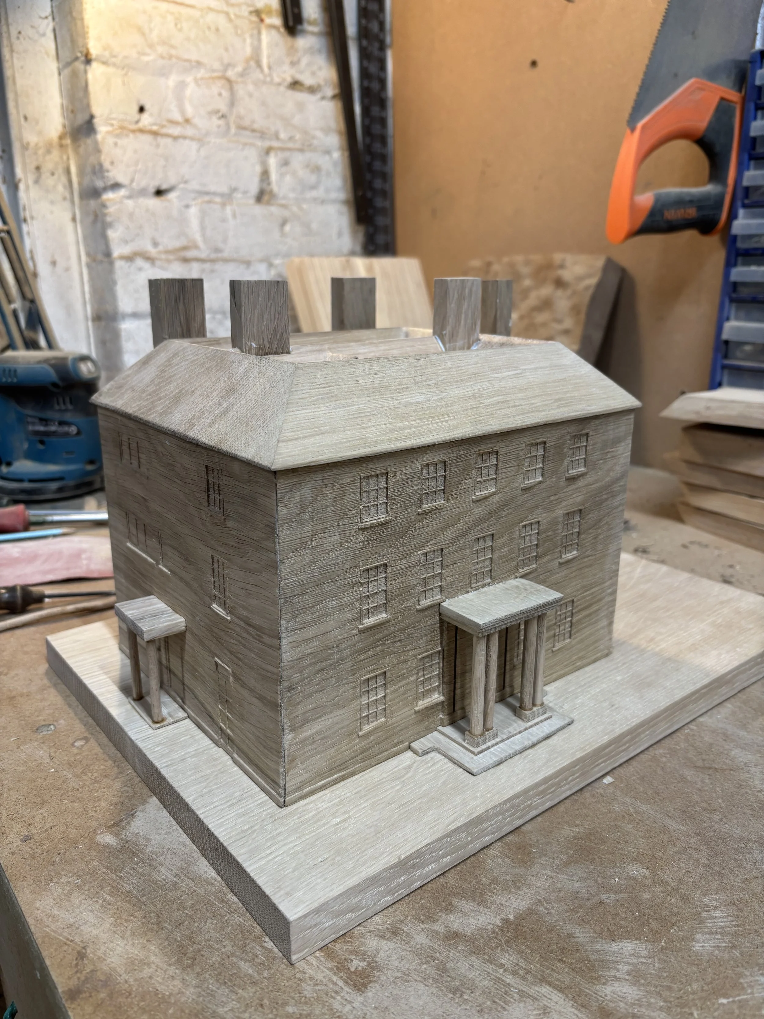 Wooden model of an old, multi-story building with a rectangular shape and classic architecture, featuring four small columns at the front entrance and multiple windows, placed on a wooden base in a workshop setting.