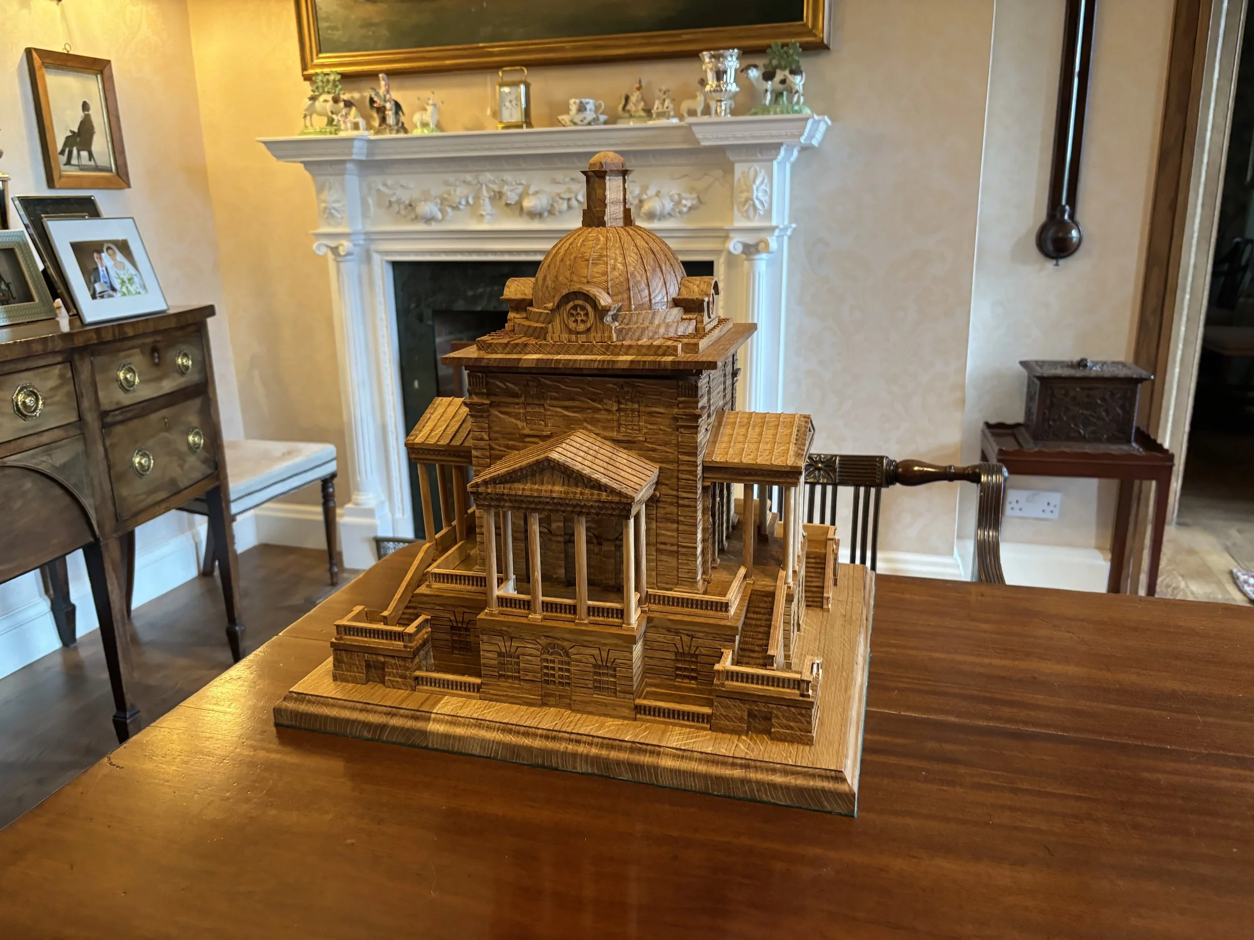 A detailed wooden model of a classical building or temple with a central dome, columns, and stairs, placed on a wooden table in a room with elegant furniture and decor.