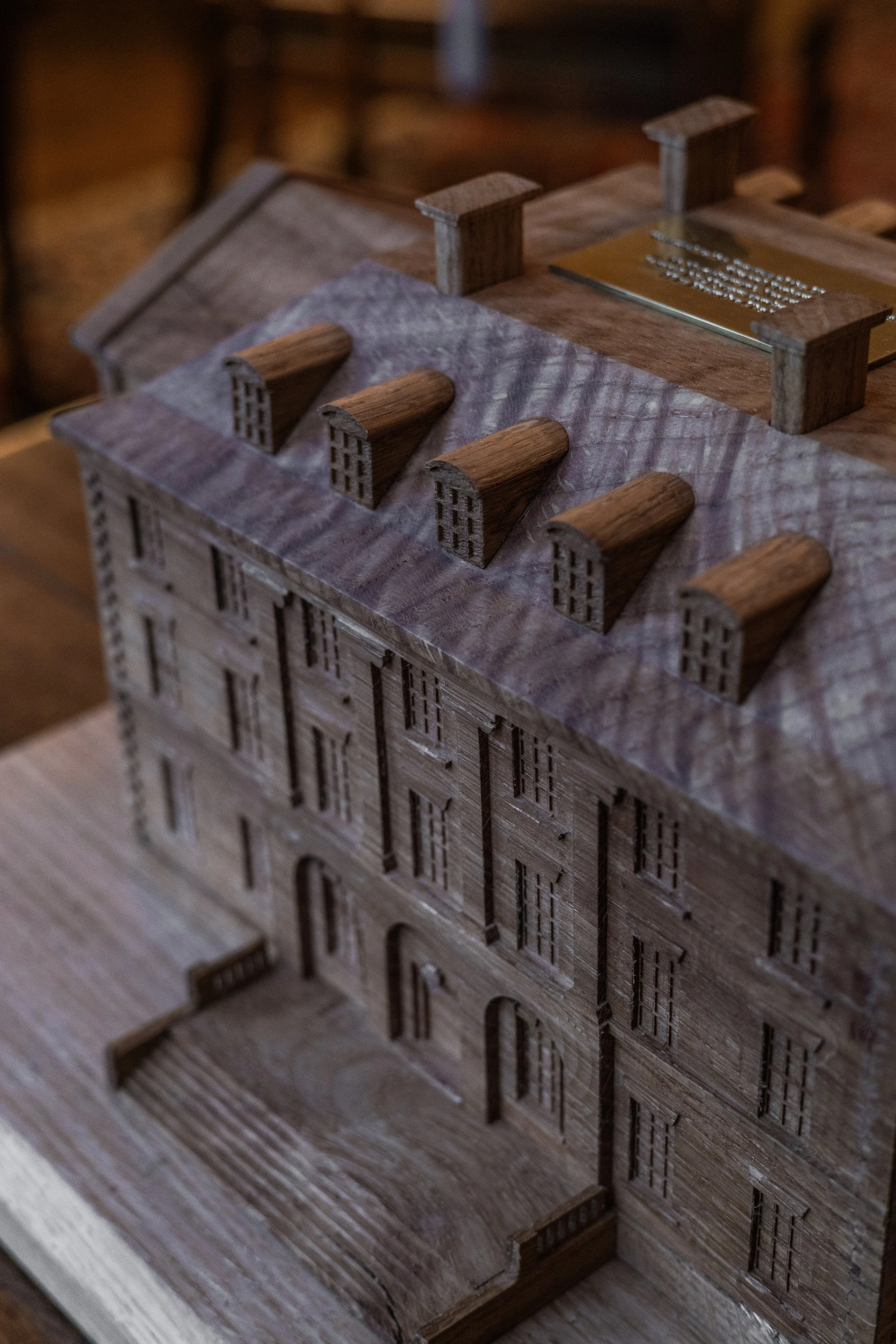 Wooden architectural model of a multi-story building with detailed windows and a front door, features a small balcony or terrace at the entrance, situated on a wooden base.