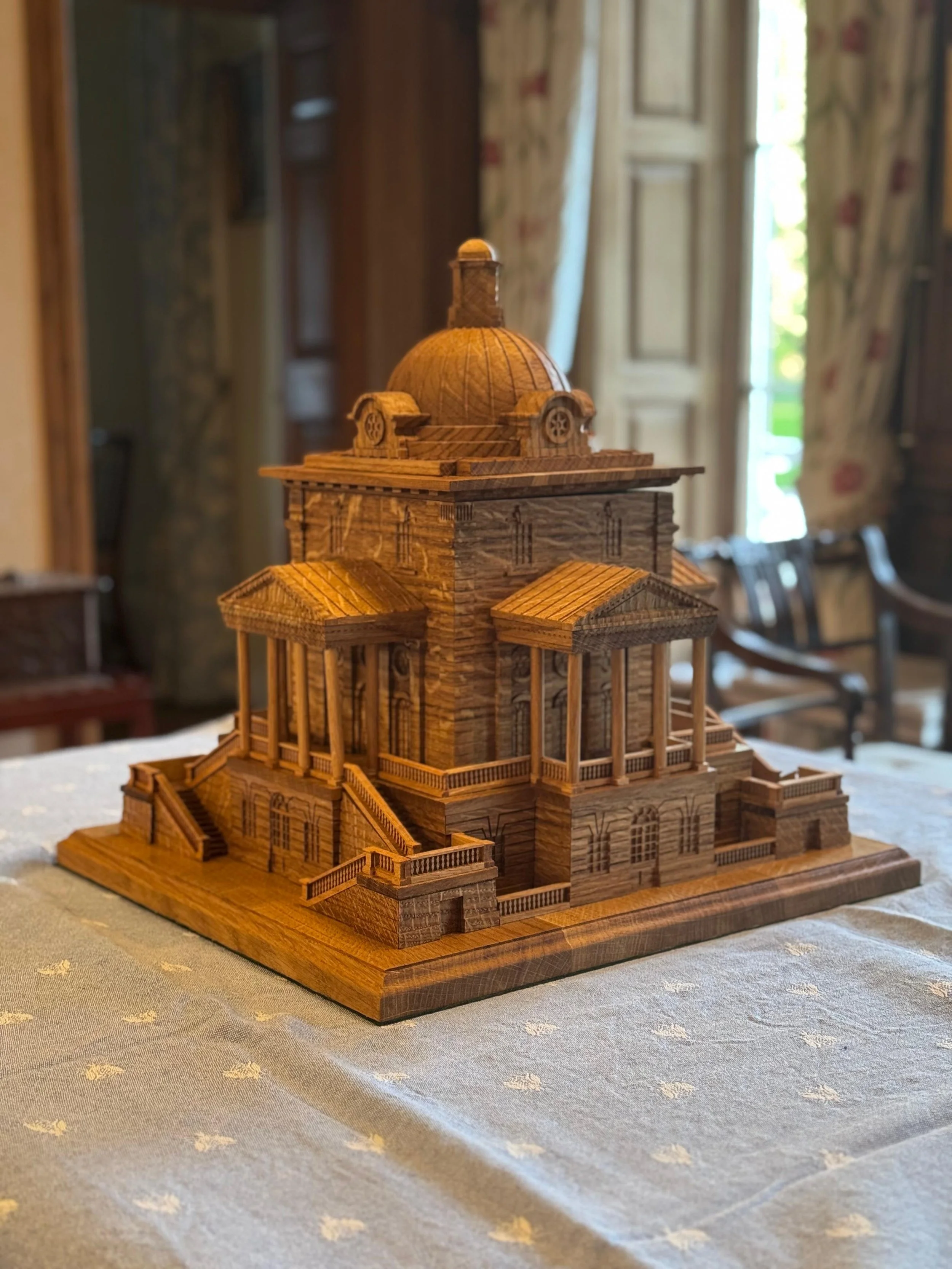 A detailed wooden architectural model of a historic building with a domed roof, steps, and columns, placed on a table in a room with wooden furniture and patterned curtains.