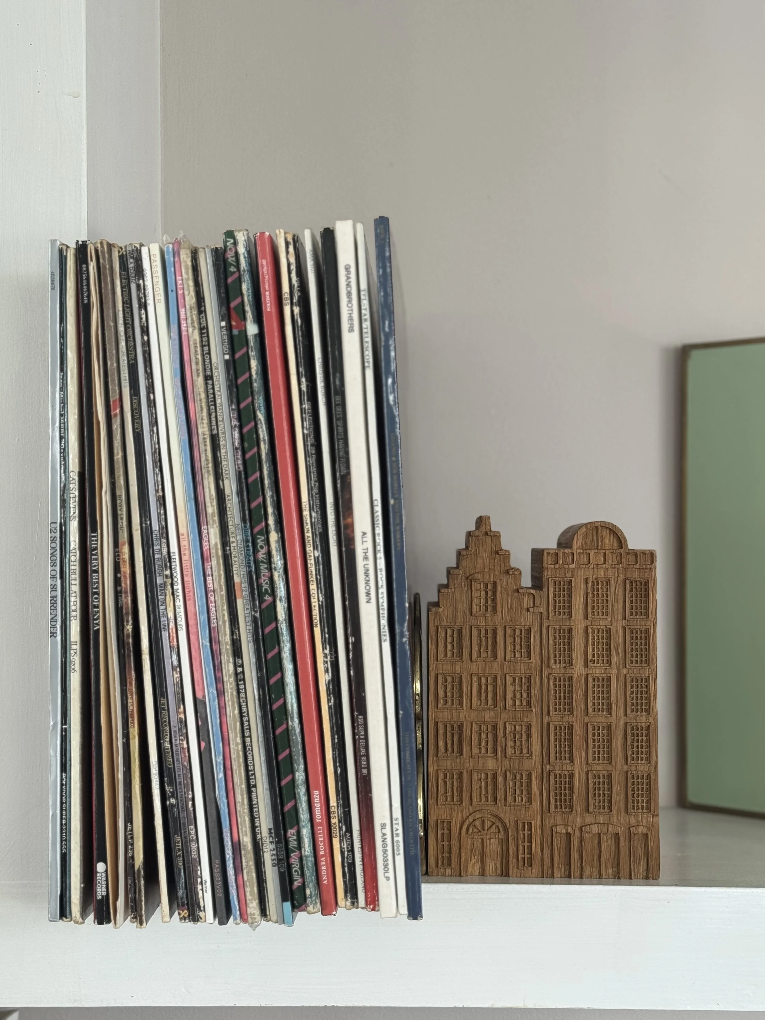 A row of vinyl records on a white shelf, next to a wooden decorative piece shaped like a building.