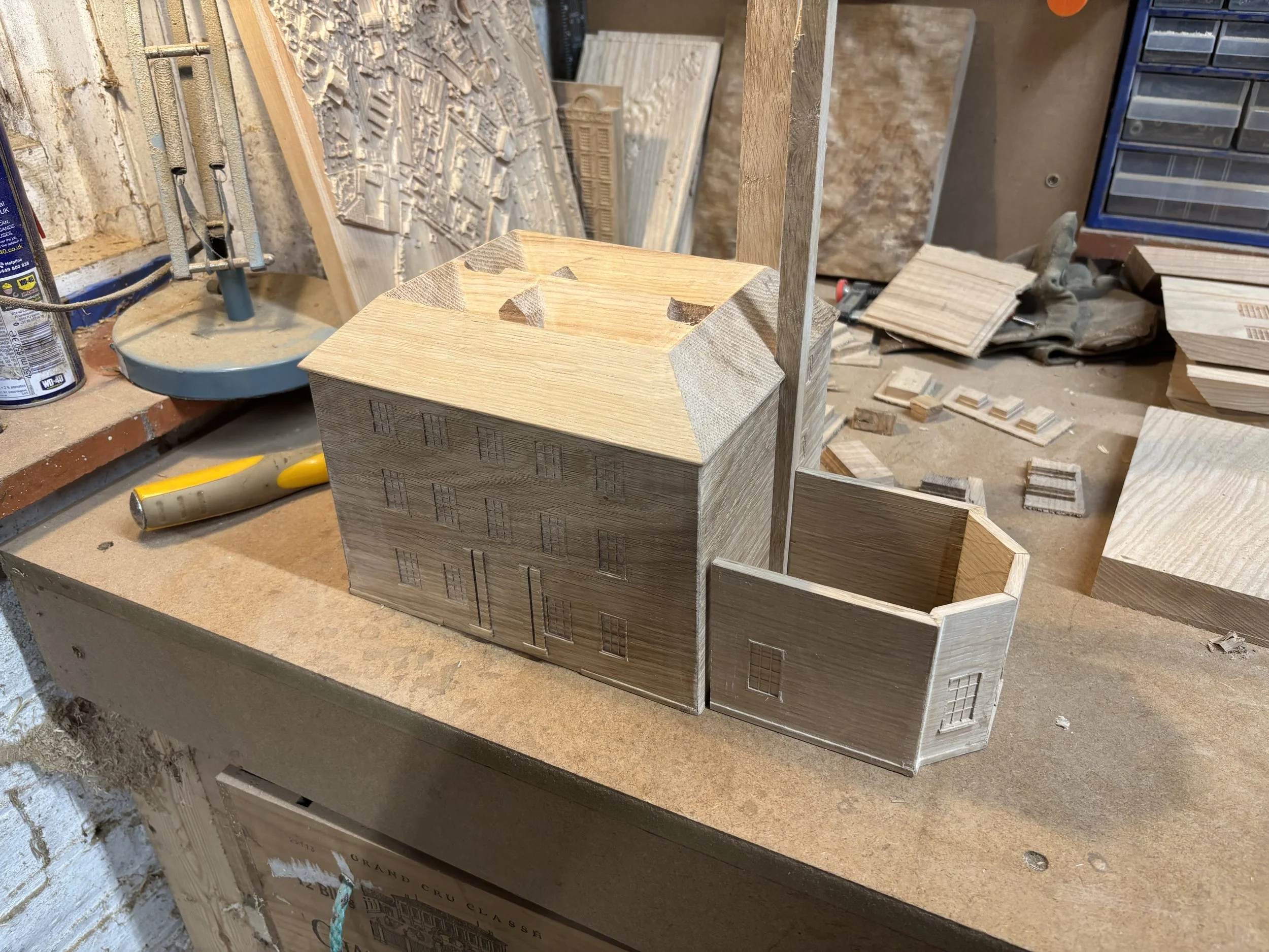 Wooden architectural model of a building with multiple levels and a small square extension, placed on a workbench with tools and other wood pieces in the background.