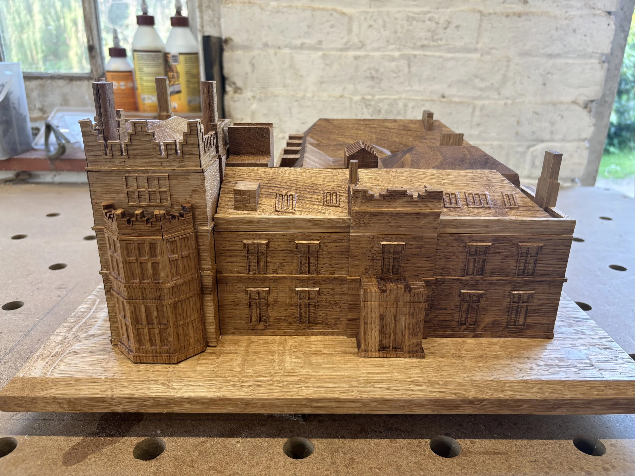 A detailed wooden model of a multi-story building with architectural features such as a tower with battlements on the left, various windows with carved outlines, and a stepped roof design, placed on a workbench in a woodworking workshop.
