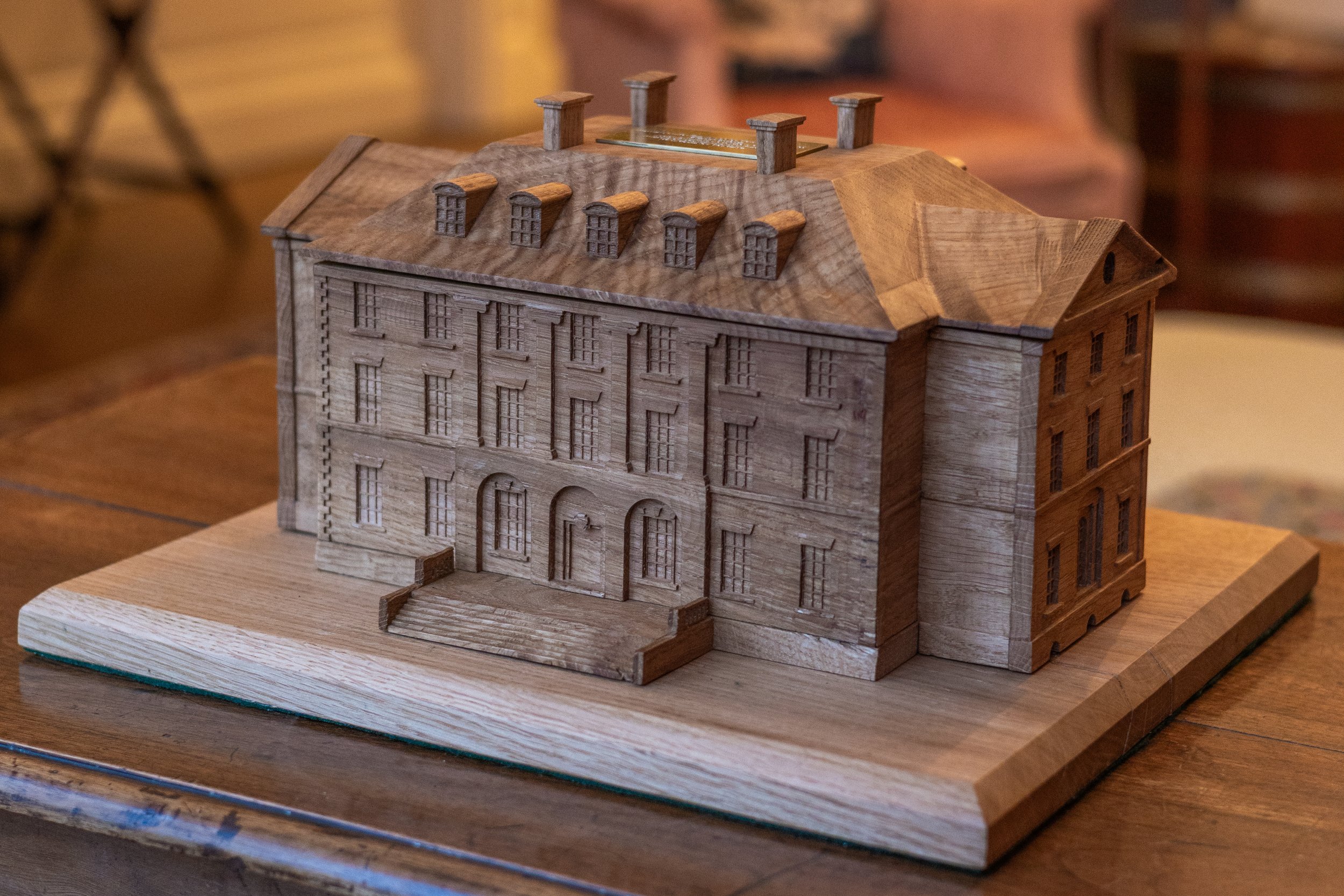 A detailed wooden model of a historic mansion or castle with multiple windows, an arched entrance, a stepped front porch, and a sloped roof with small dormer windows, placed on a wooden base.