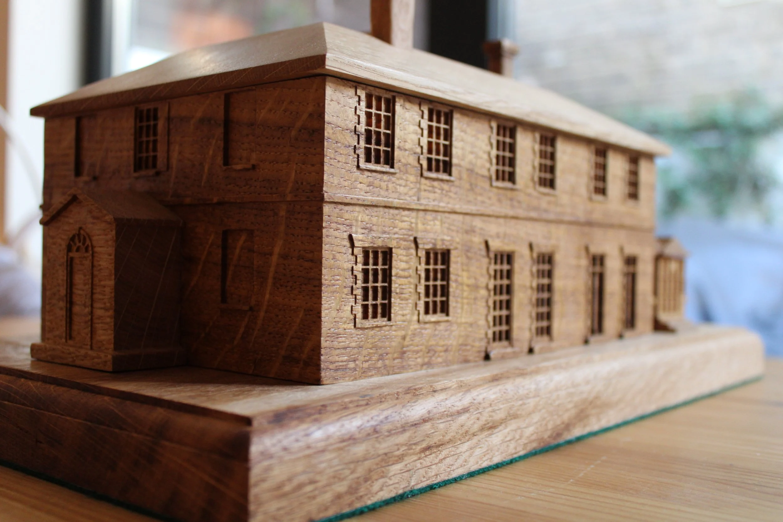 A detailed wooden model of a two-story brick house with multiple windows, a small porch, and a sloped roof on a wooden base.