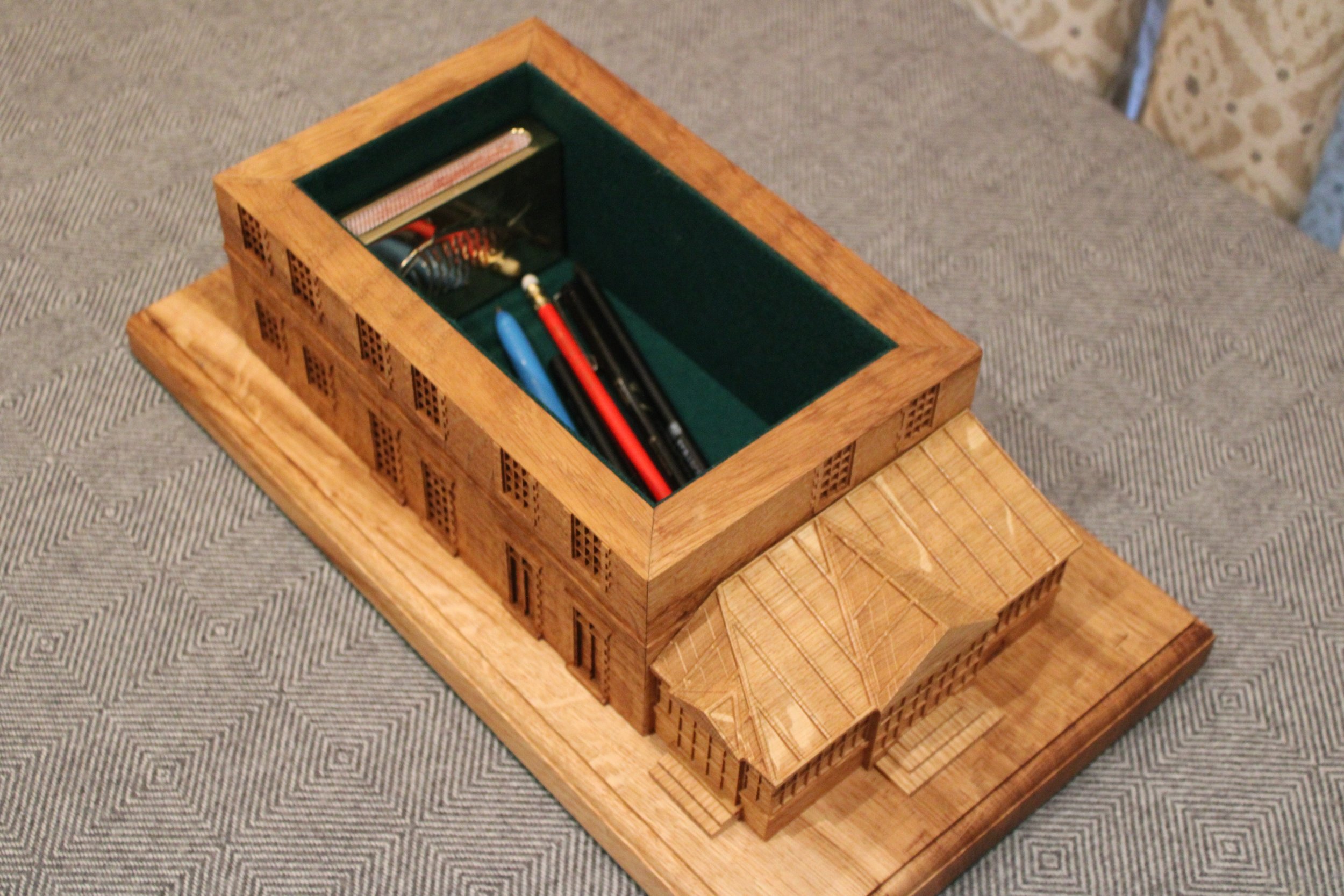 A wooden model of a building with a green interior, placed on a wooden base, with various miniature objects inside.
