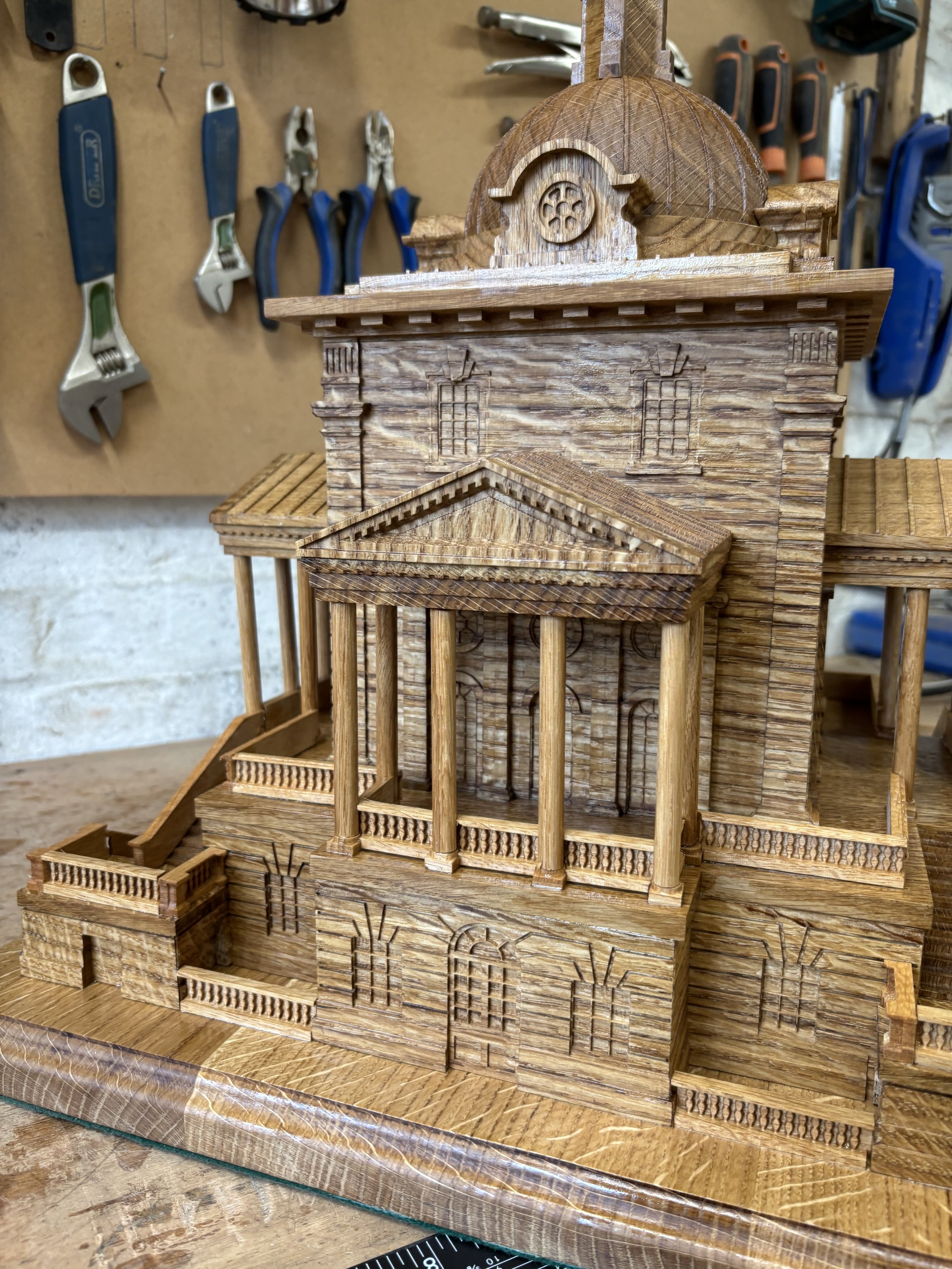 Wooden architectural model of a classical building with columns and a domed roof, set on a workbench with woodworking tools hanging in the background.