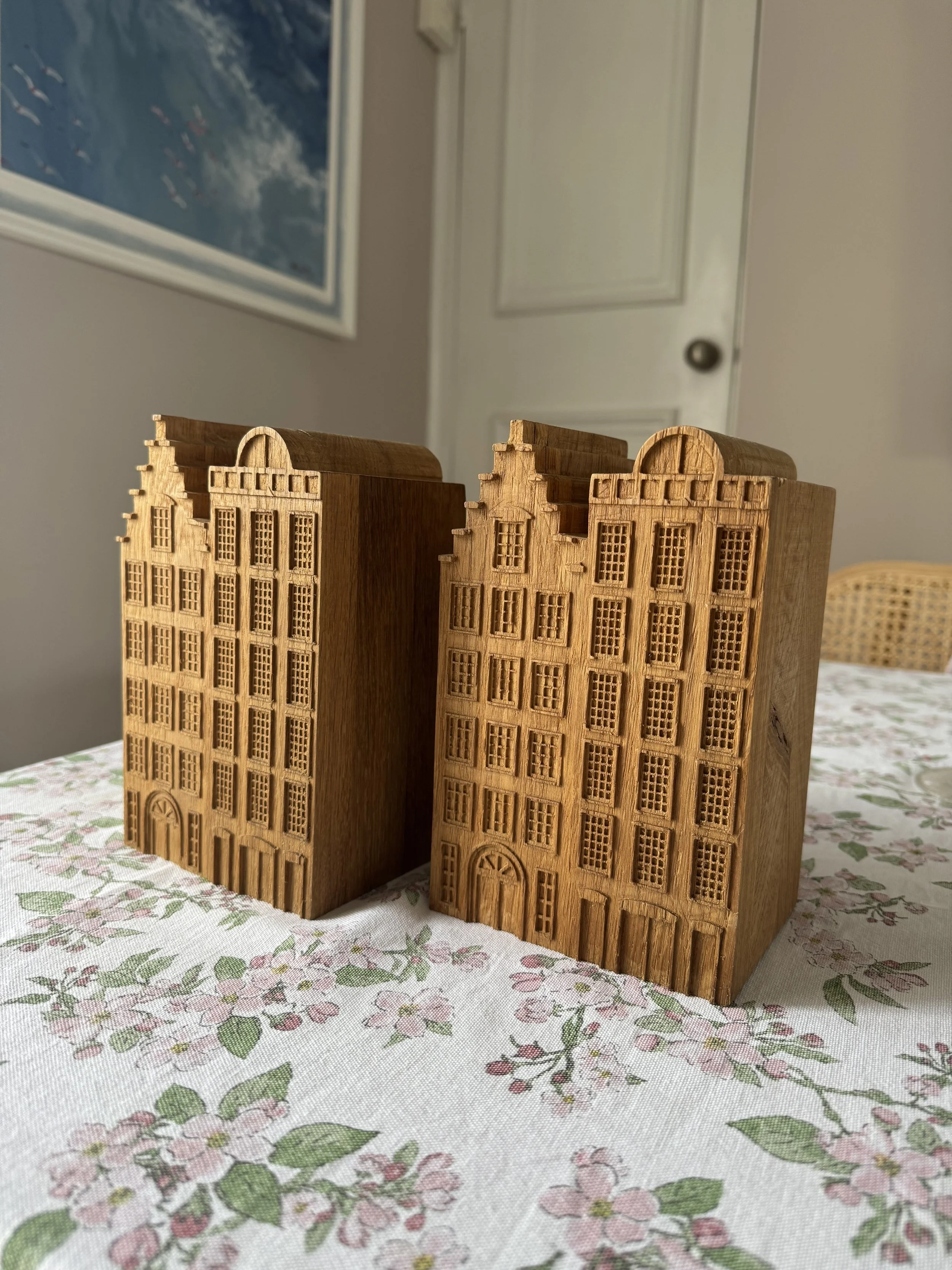 Two wooden miniature building models with detailed architectural features on a floral tablecloth, with a door and multiple small windows, set in a room with a door and a painting in the background.