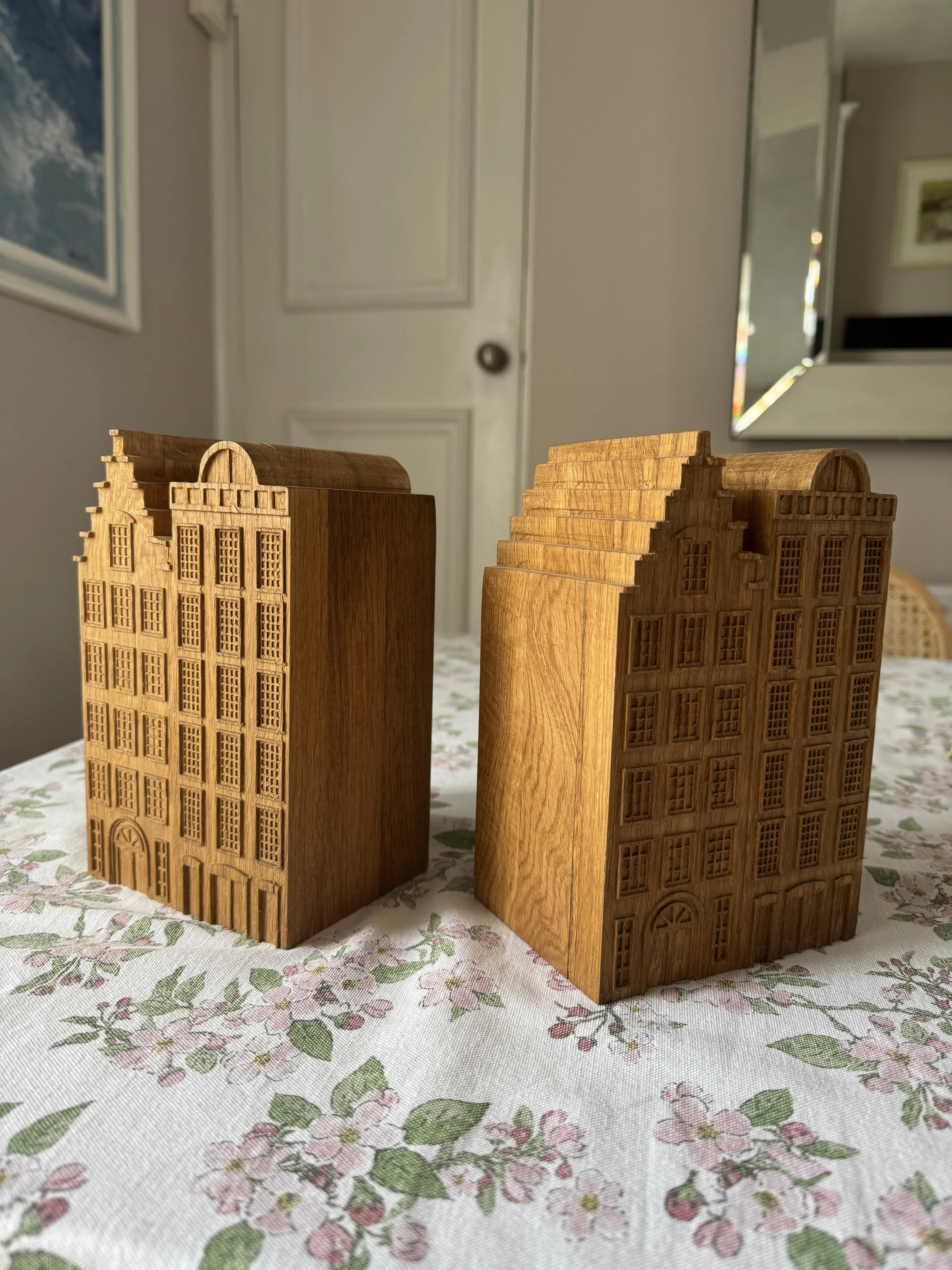 Two wooden miniature buildings with detailed windows and doors, placed on a floral-patterned tablecloth in a room with a door and mirror in the background.