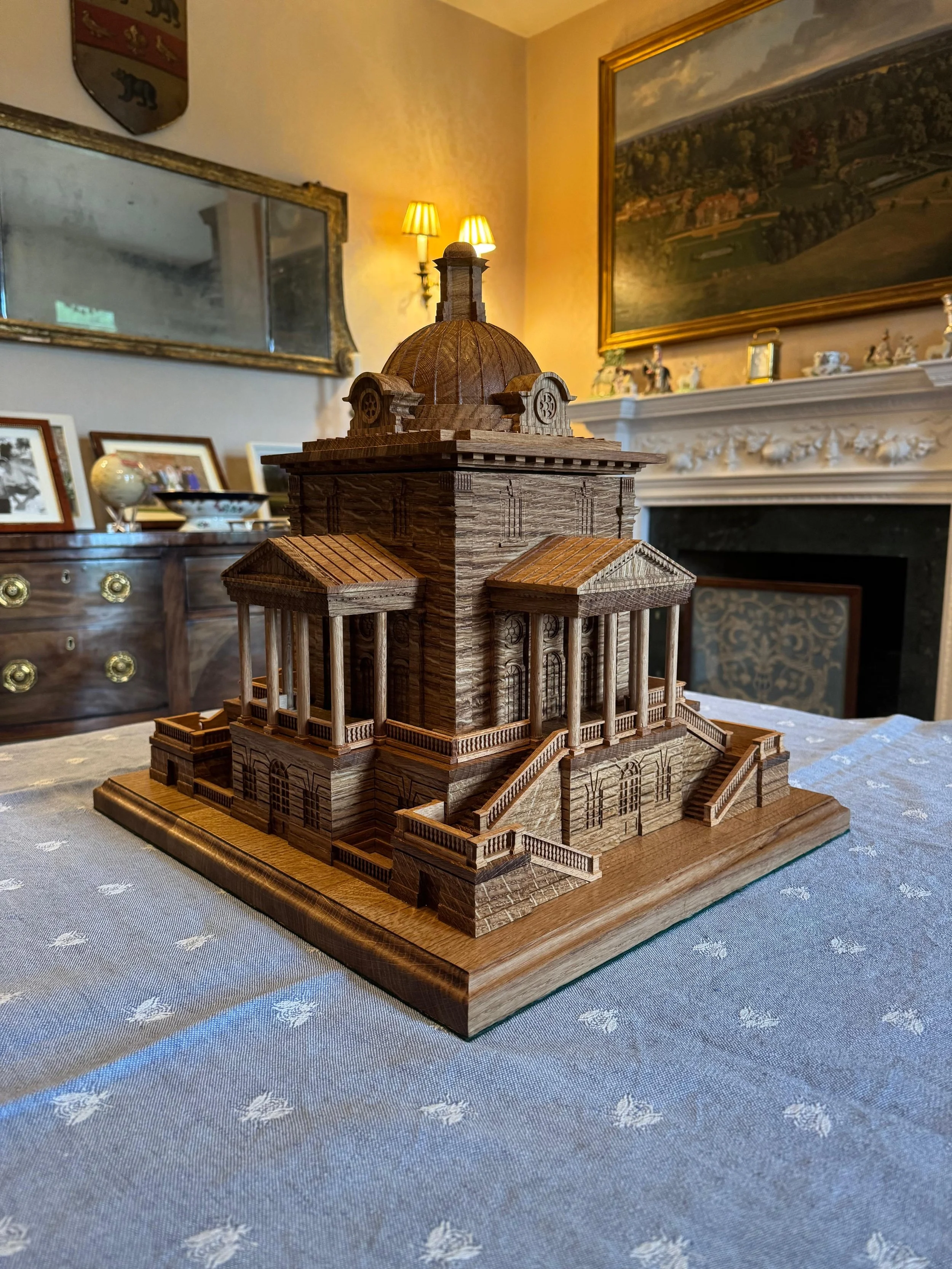 A detailed wooden model of a large, ornate building with a dome, stairs, and multiple columns, sitting on a table in a decorated room.