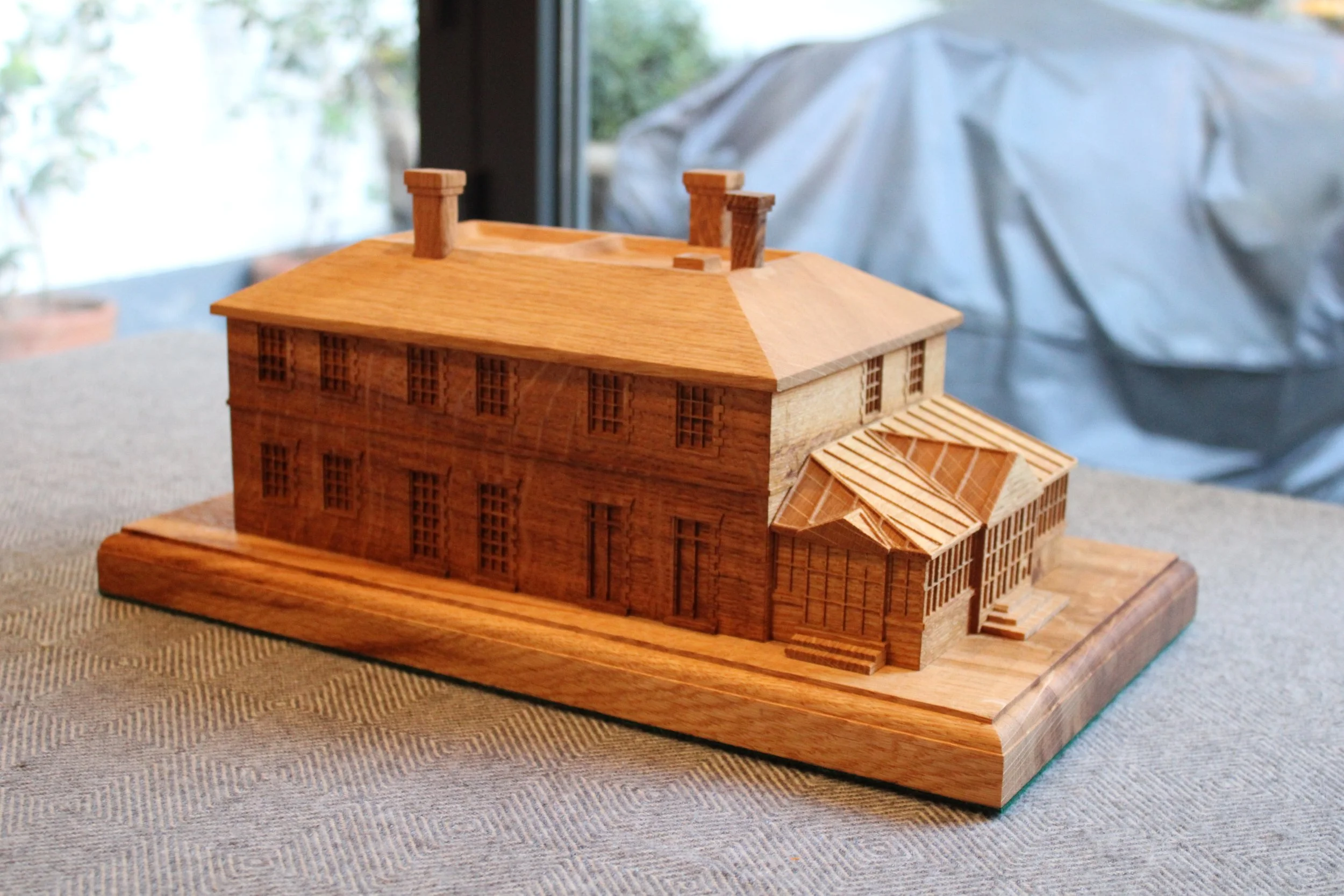 Wooden architectural model of a multi-story building with a sloped roof and chimney, placed on a wooden base.