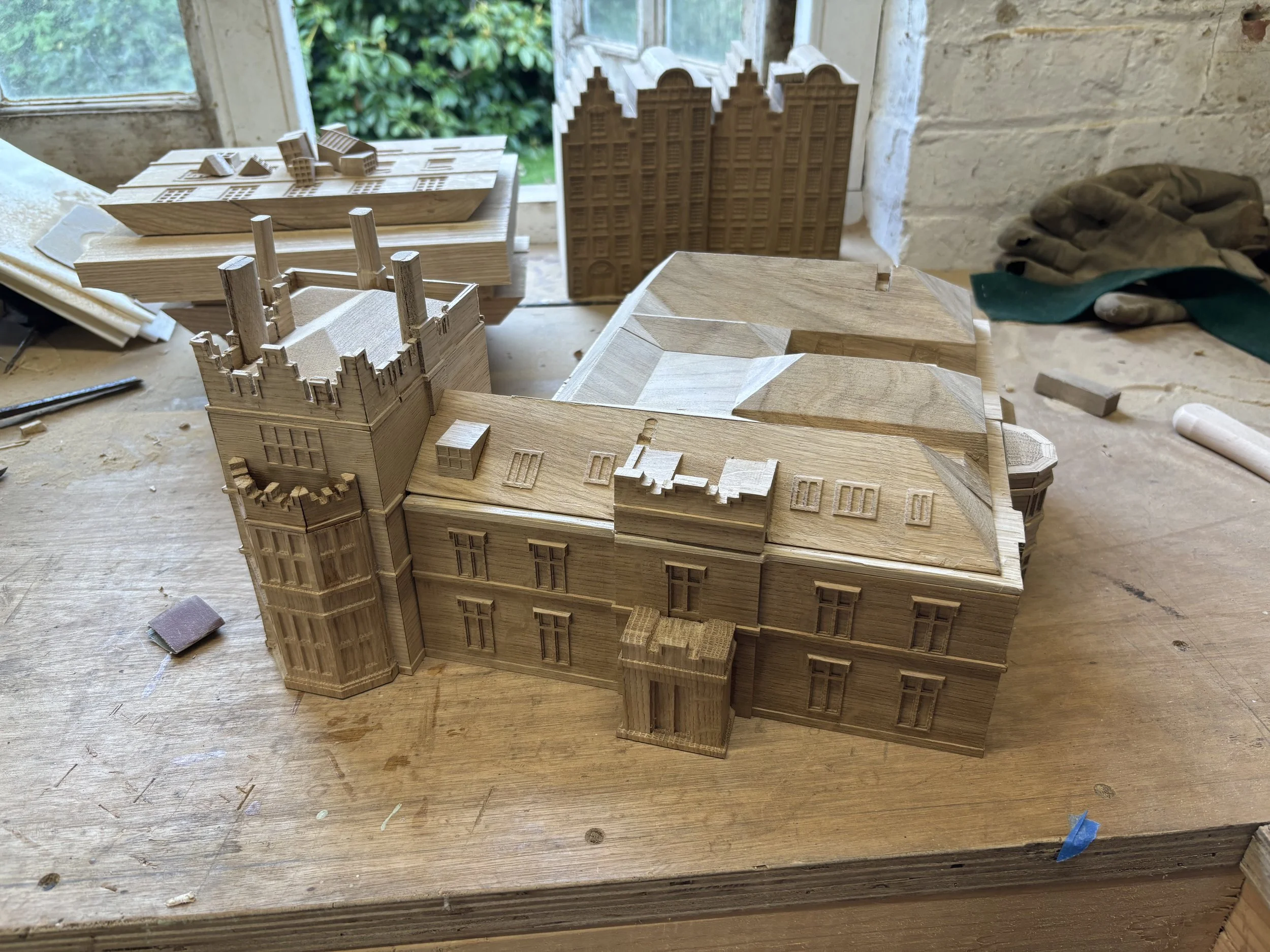 A detailed wooden architectural model of a castle and other buildings on a workbench in a workshop.