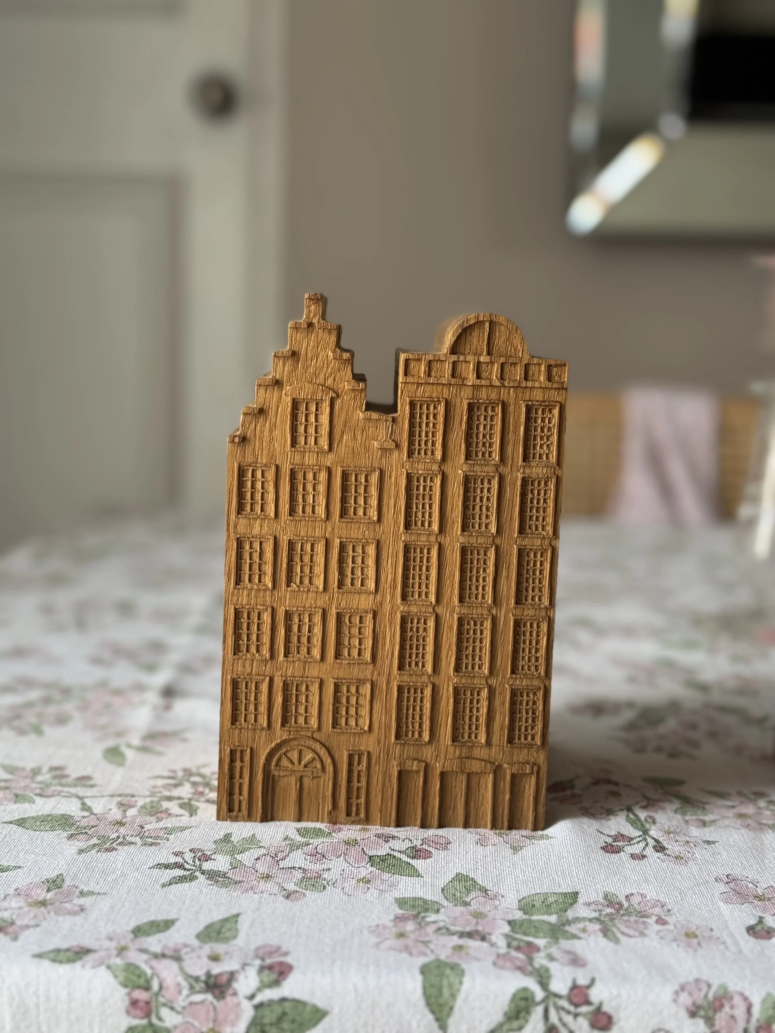 A small wooden carving of a tall, narrow building with multiple windows and a door, placed on a floral tablecloth in a room.