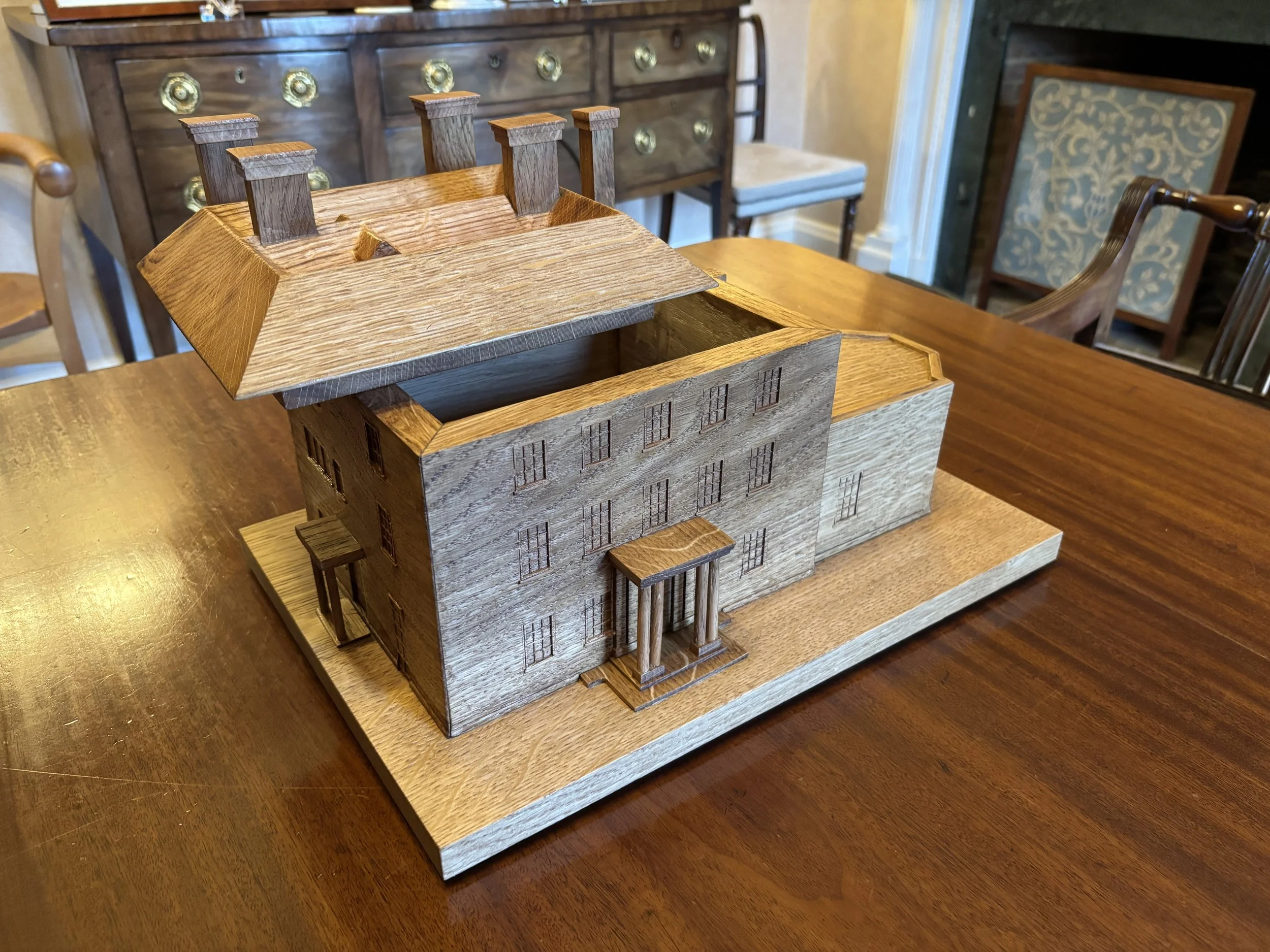 A wooden architectural model of a house with a pitched roof, multiple windows, a small front porch, and a chimney, placed on a wooden table.