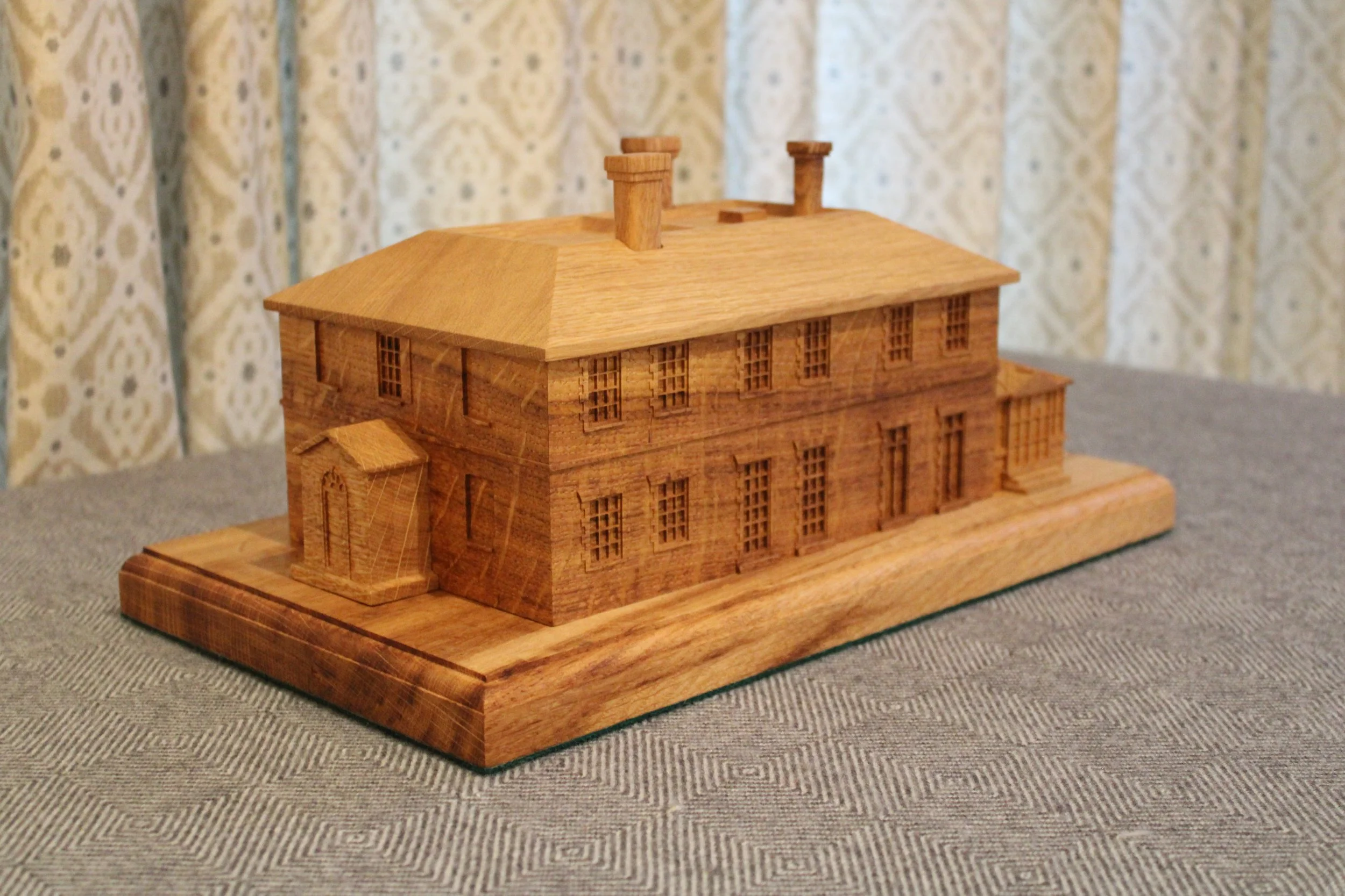 A detailed wooden model of a historic two-story building with multiple windows, a sloped roof with chimneys, and a side door, placed on a wooden base on a gray fabric surface.