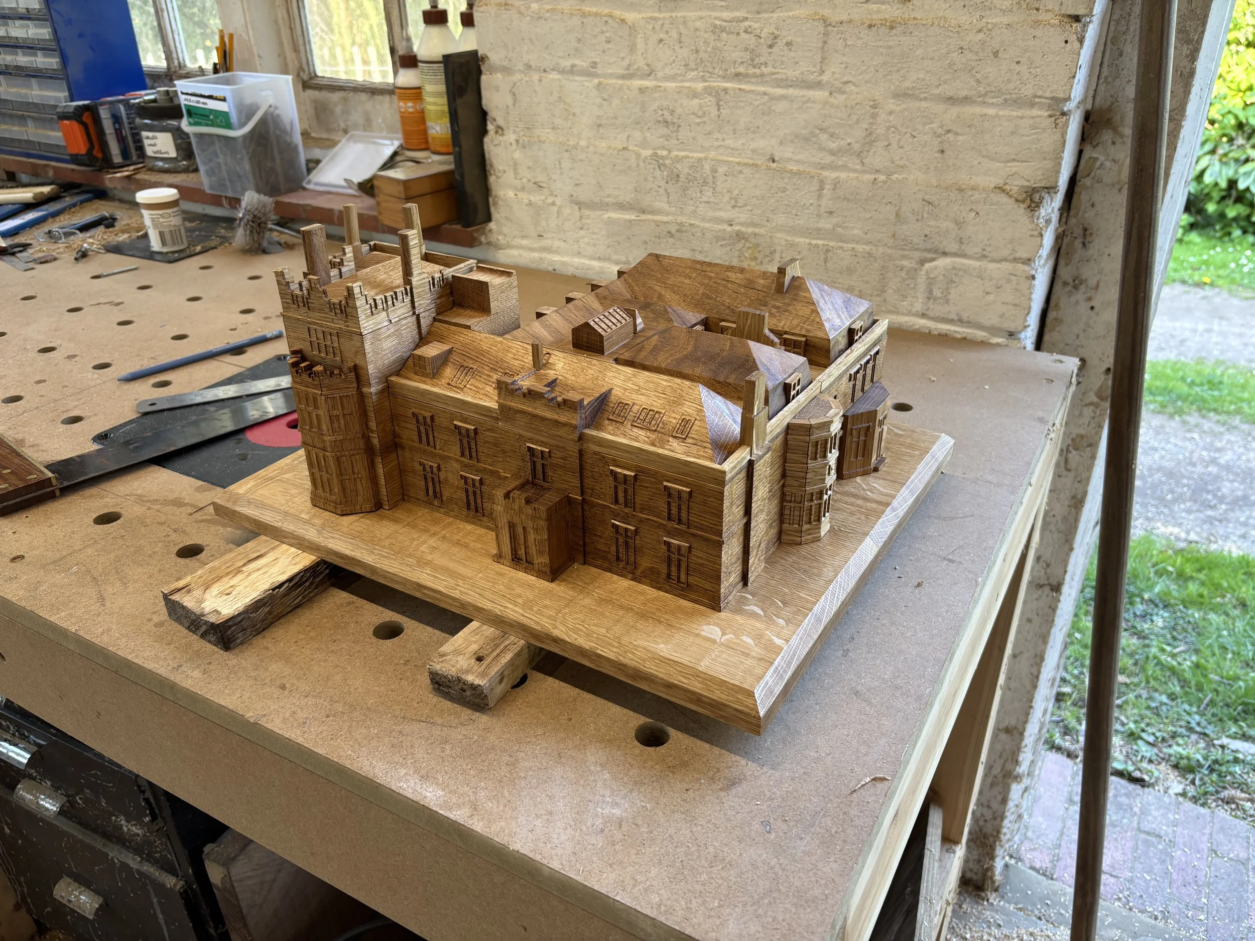 A detailed wooden model of a castle with towers and walls, situated on a workbench in a workshop.