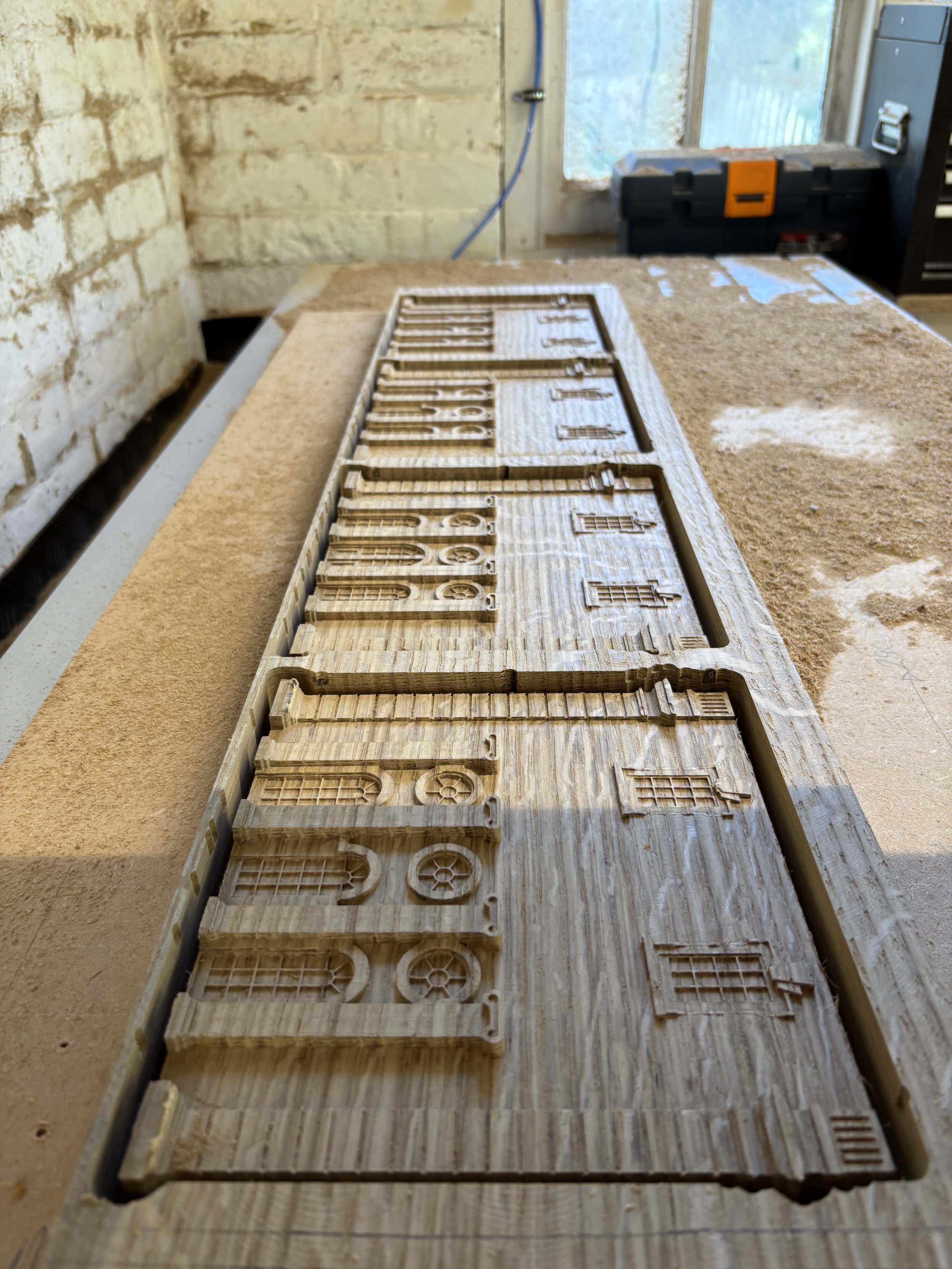 A wooden mold or stamp with engraved buildings, placed on a wooden work surface in a workshop. The background shows a window, a brick wall, and some workshop tools.
