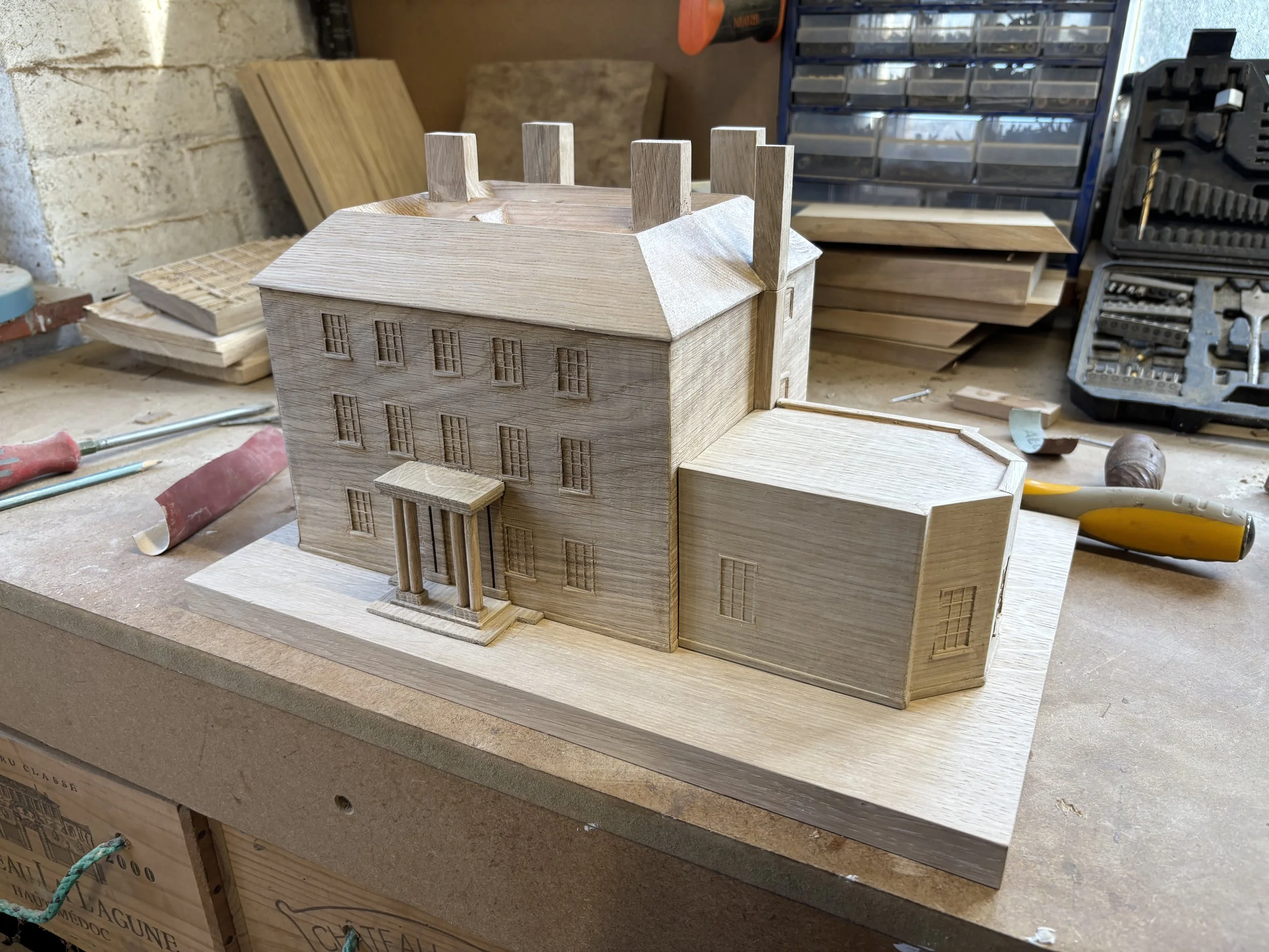 Wooden architectural model of a house in a workshop, with tools and wood pieces in the background.
