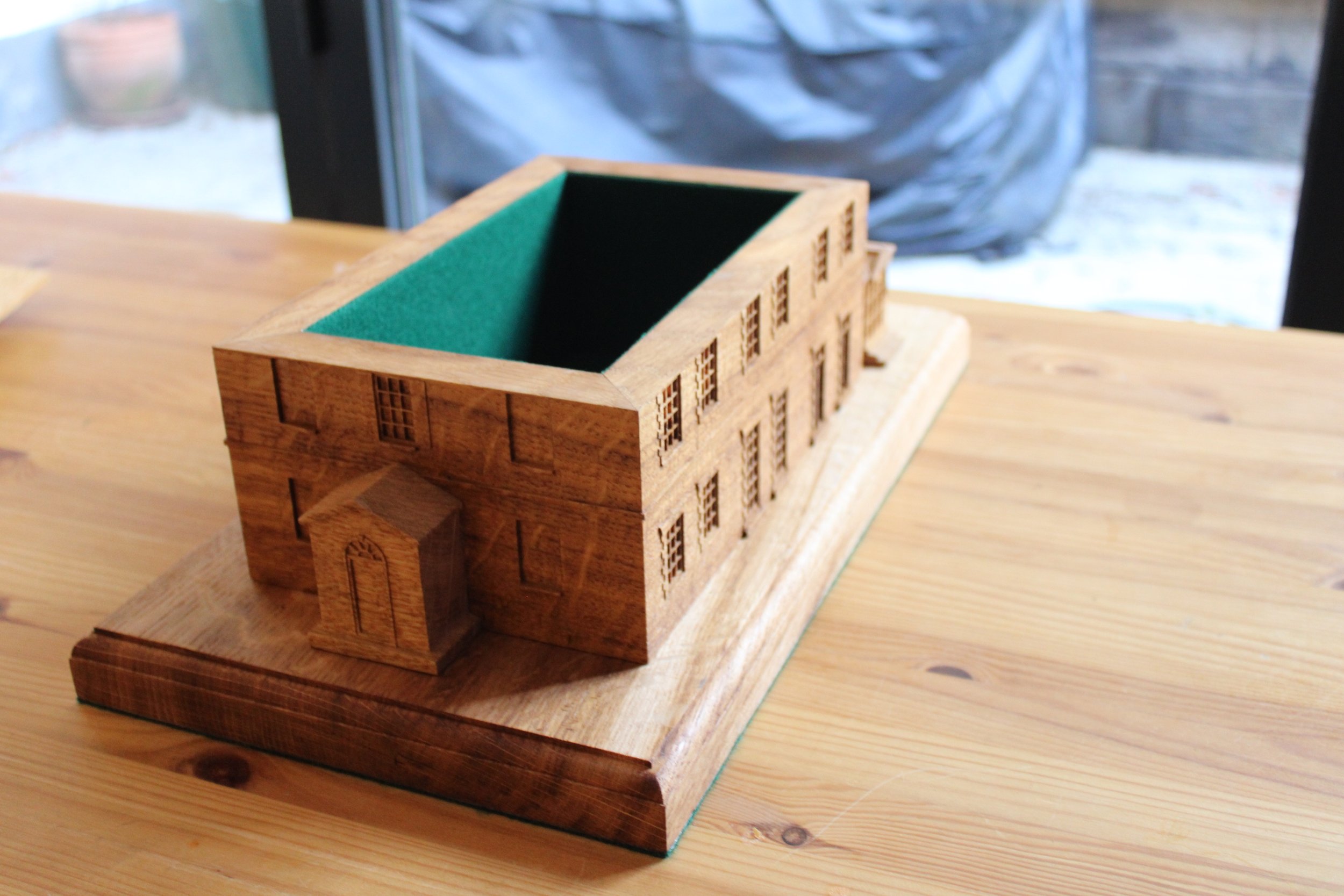 A miniature wooden model of a building with a green felt interior, placed on a wooden table.