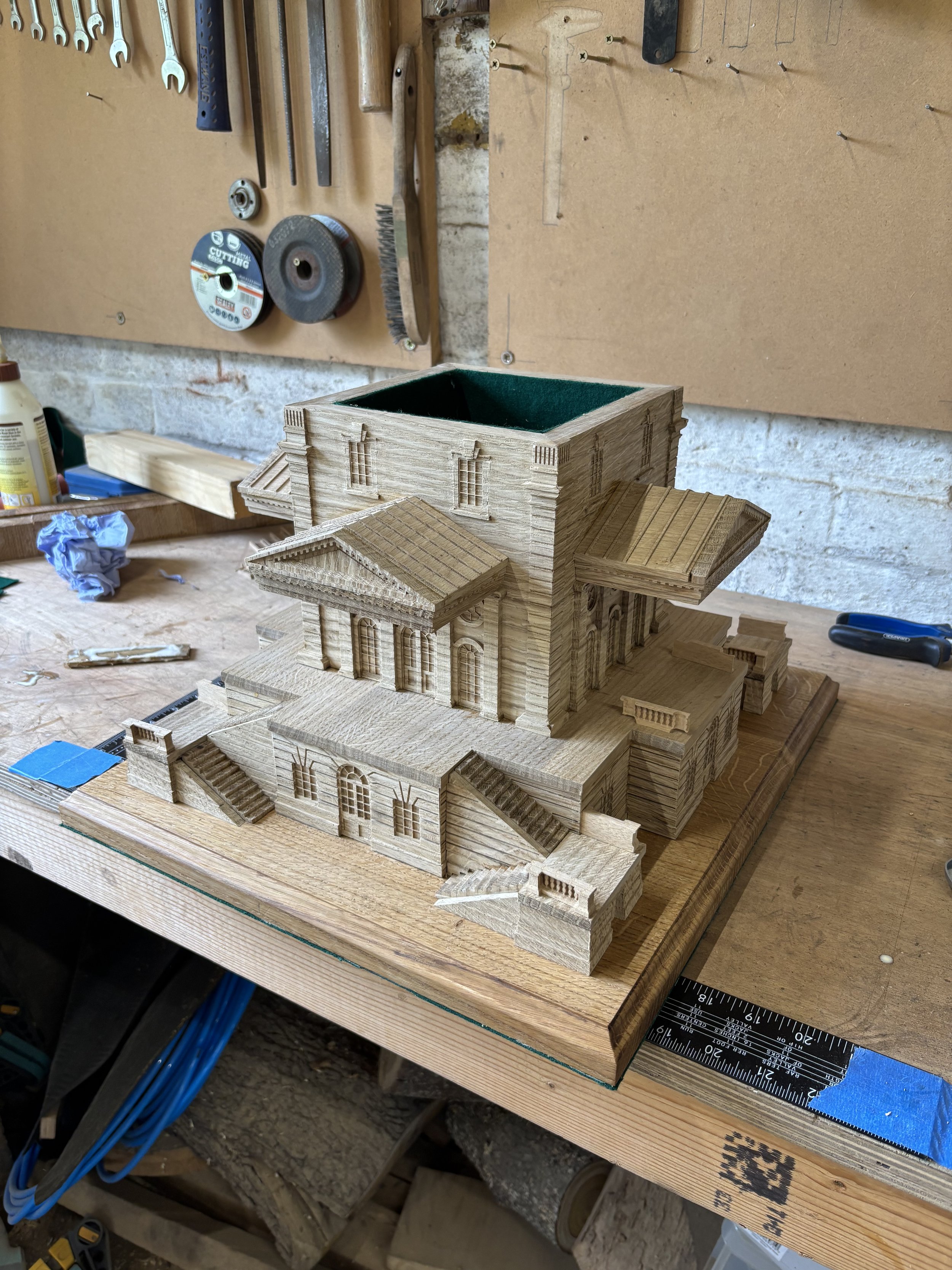 A detailed wooden architectural model of a multi-story building with stairs, columns, and windows, placed on a workbench in a woodworking shop.