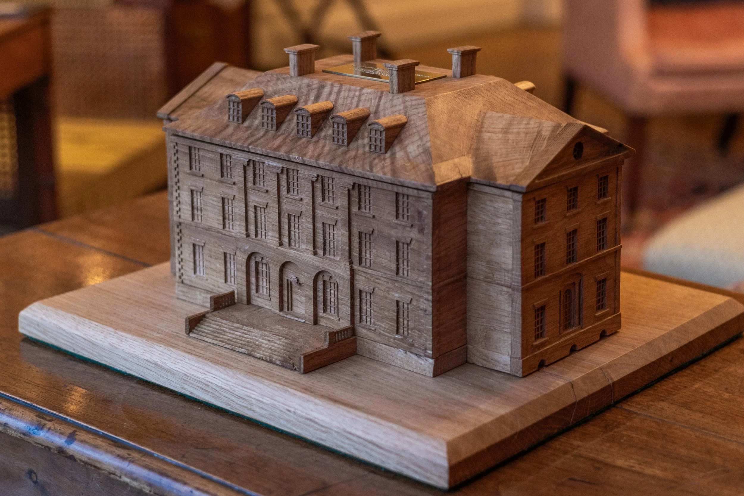 A small wooden model of a four-story building with a pitched roof, multiple windows, and a staircase leading to the front entrance, placed on a wooden table.