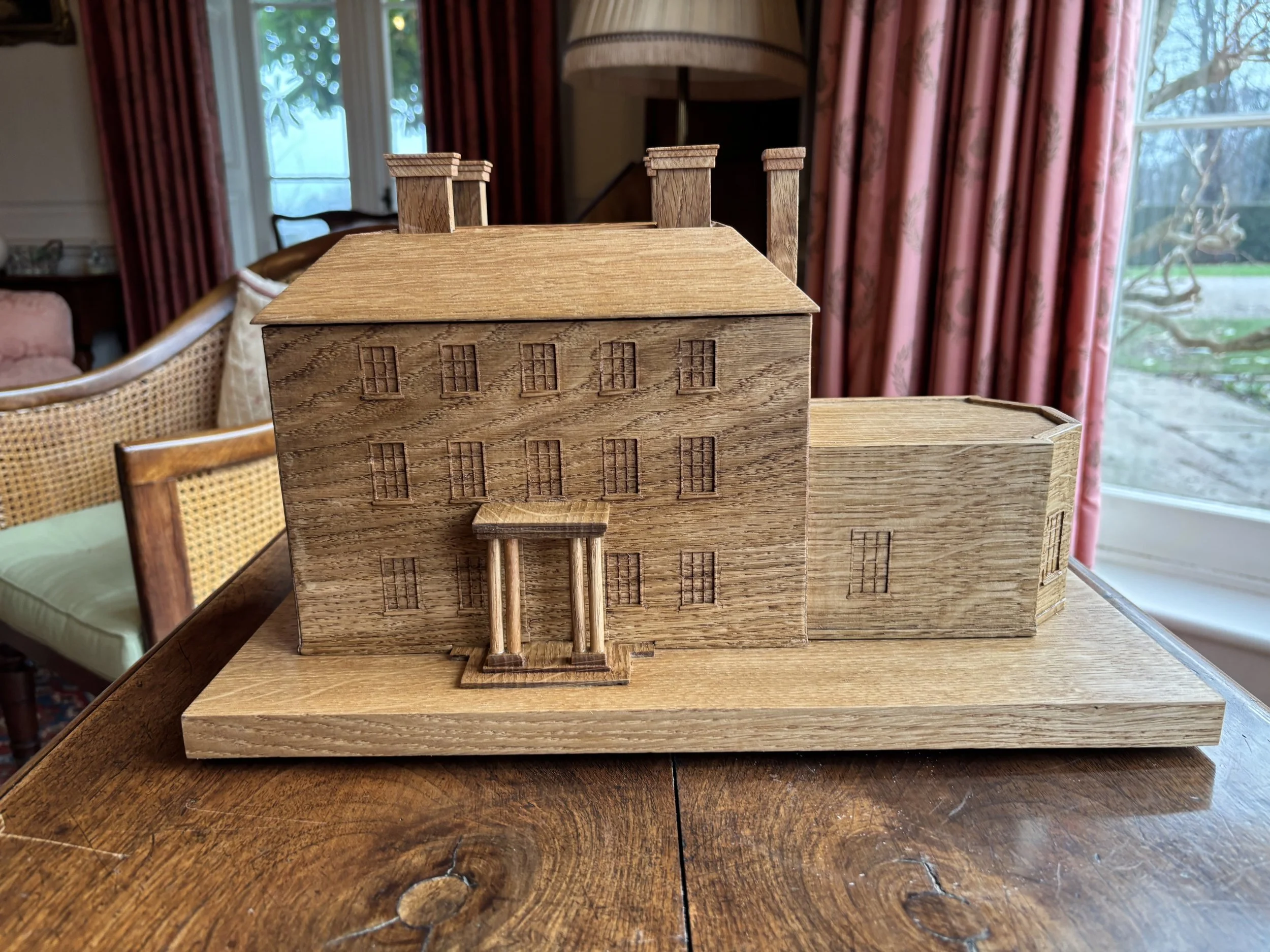 A detailed wooden model of a multi-story house with a porch, chimneys, and multiple windows, placed on a wooden table inside a room with large windows and red curtains.