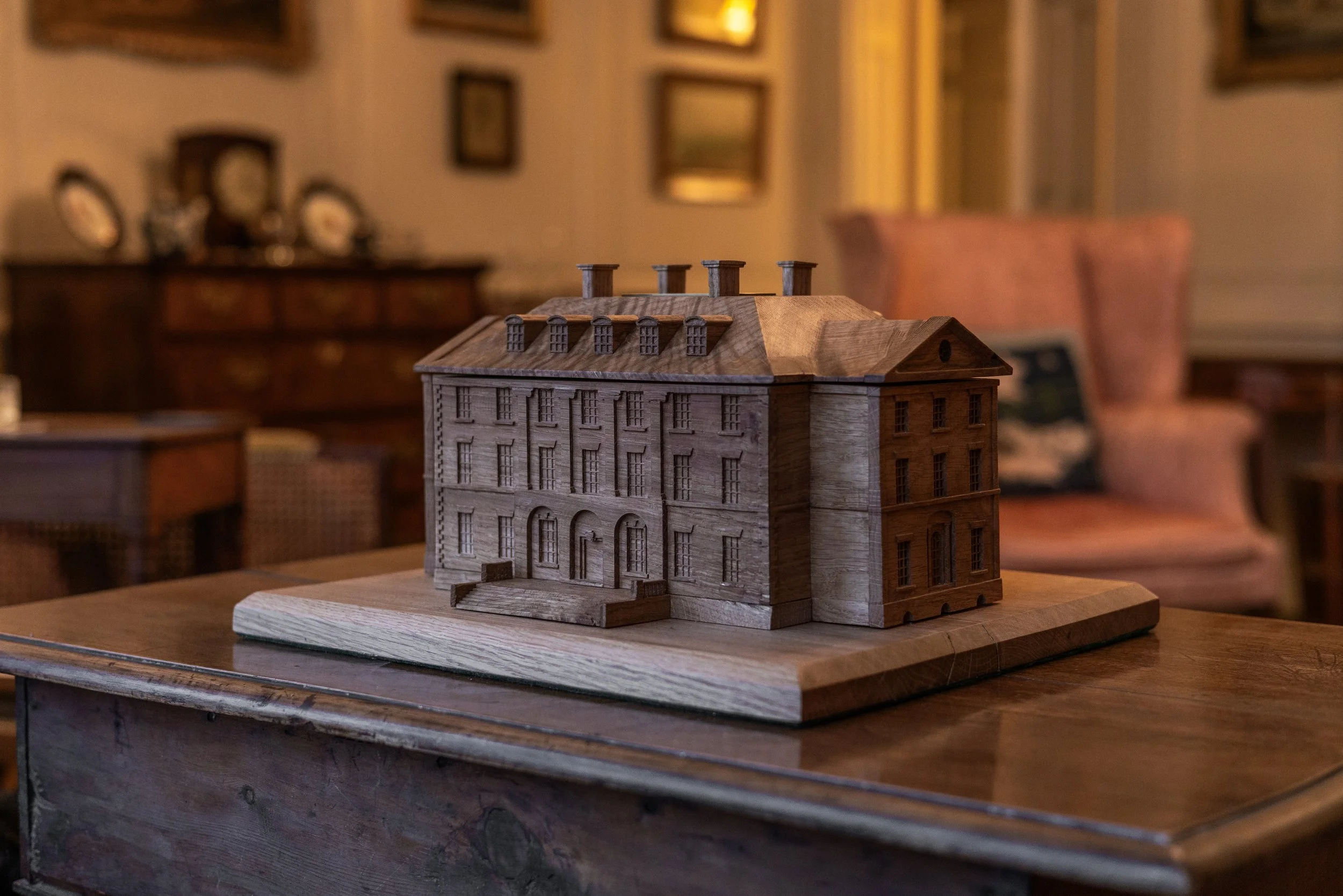 A detailed wooden model of a historic multi-story building with chimneys, placed on a wooden table in a room with vintage furniture and framed pictures on the wall.