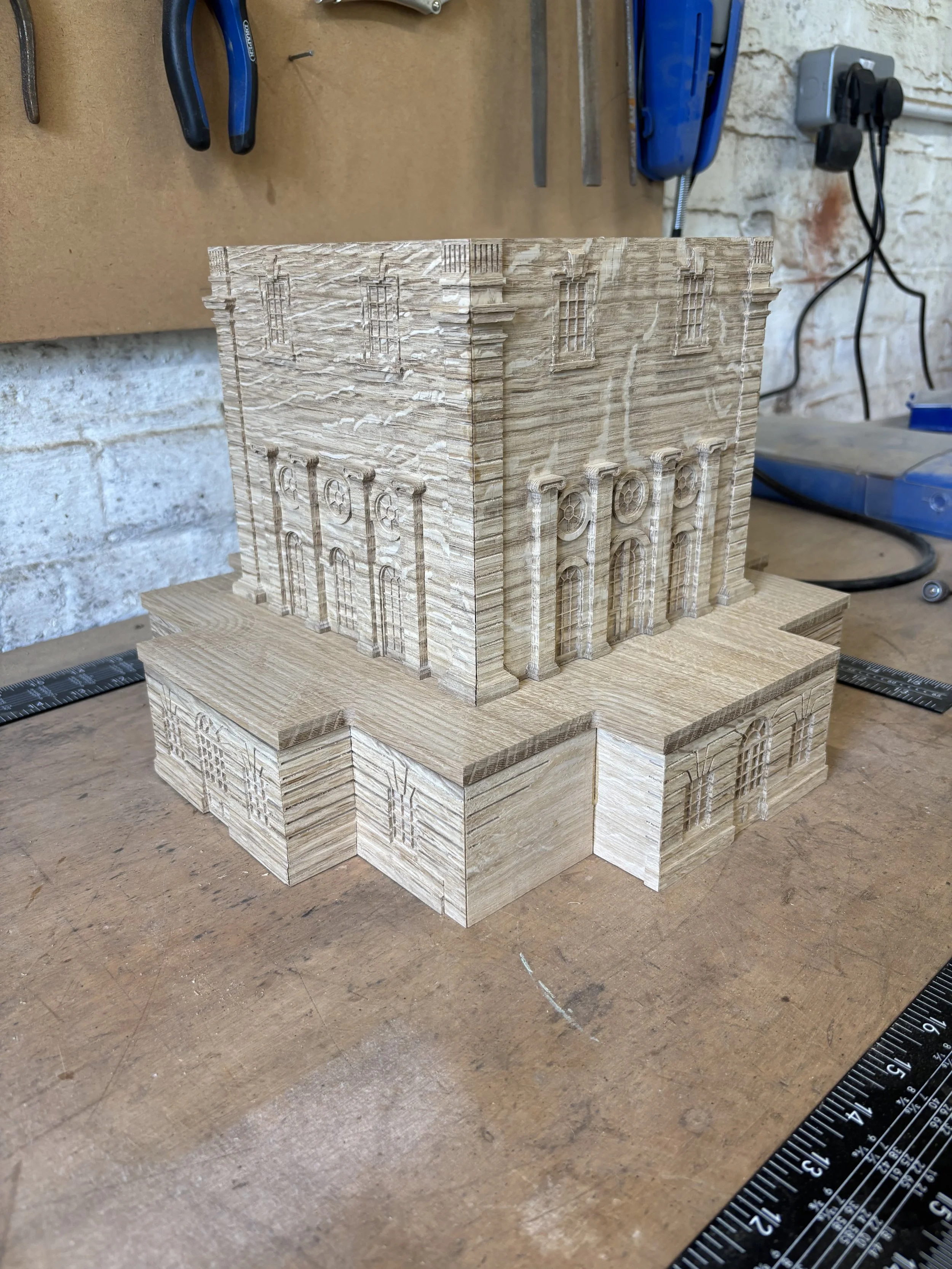 A wooden model of a building with detailed carvings of windows and architectural features, placed on a workbench in a woodworking shop.