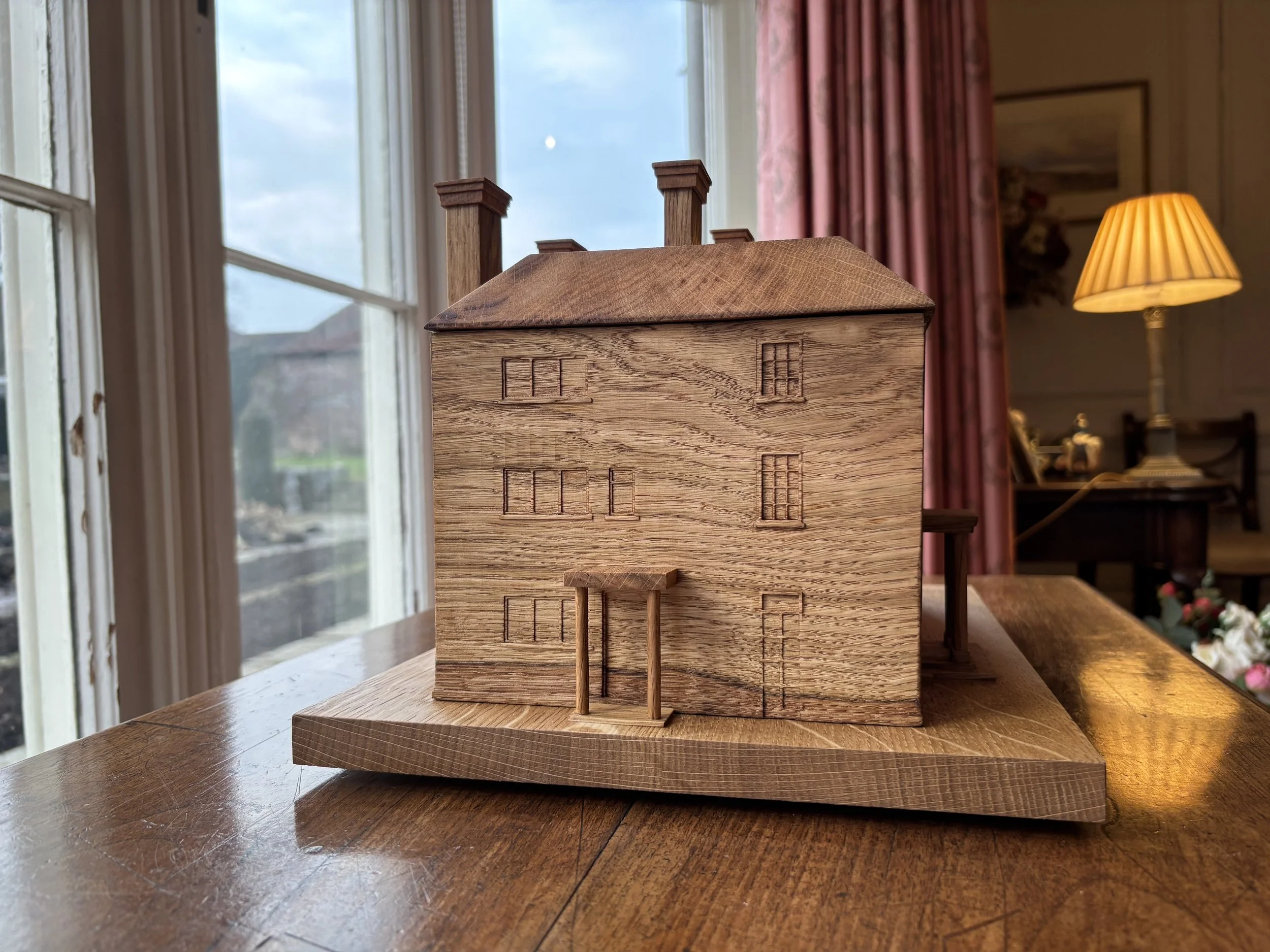 Wooden model of a house with carved windows and door, placed on a wooden table in a room with large windows and a lamp in the background.