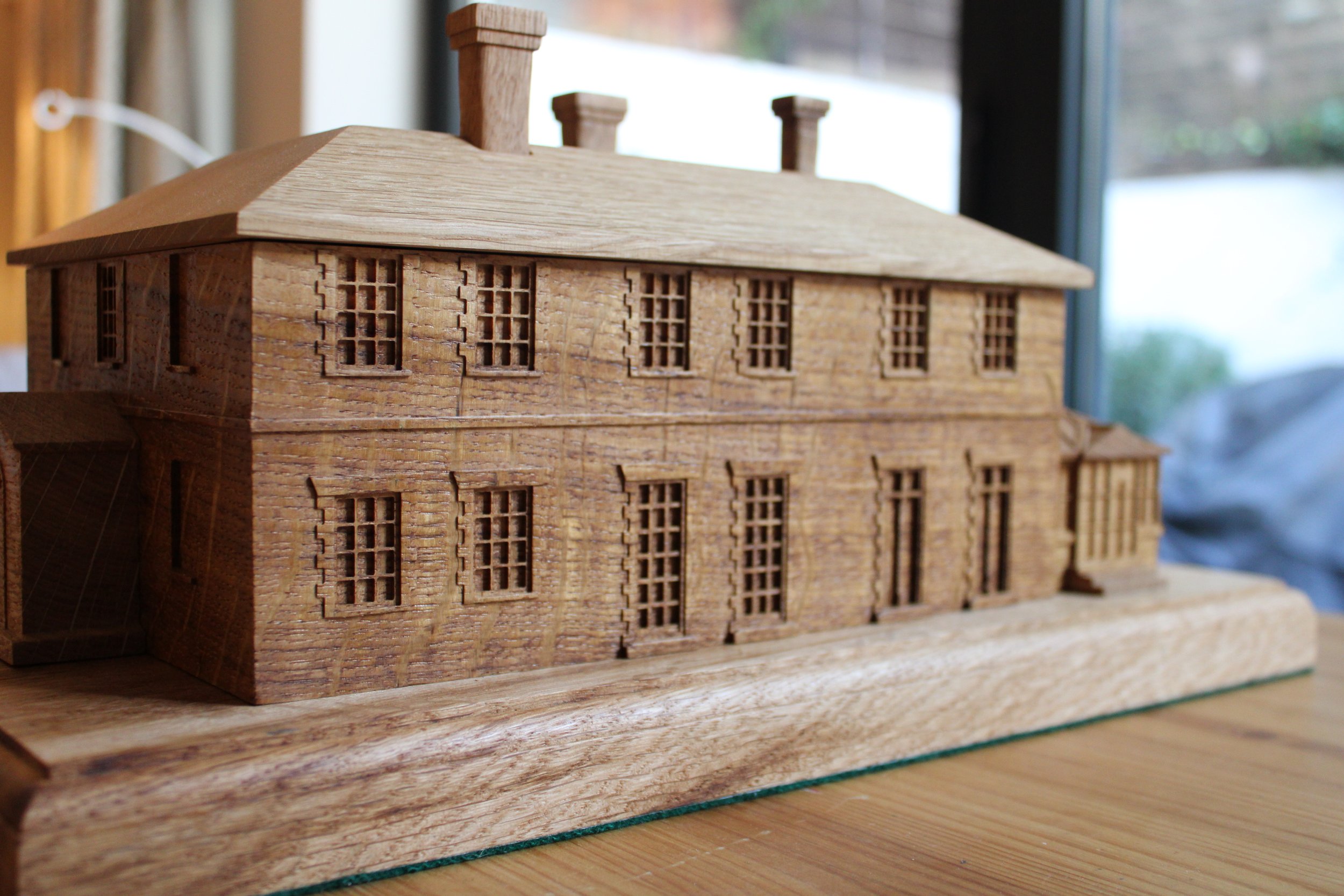 Wooden architectural model of a large building with multiple windows, chimney-like structures on the roof, placed on a wooden surface near a window.